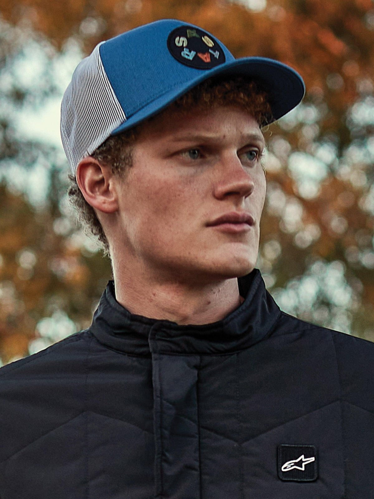 Uomo che indossa il cappello Alpinestars Gambit Trucker, berretto blu e bianco con retro in rete e patch circolare con logo Astars, abbinato a una giacca trapuntata nera Alpinestars, ritratto lifestyle casual ispirato al motorsport con sfondo autunnale