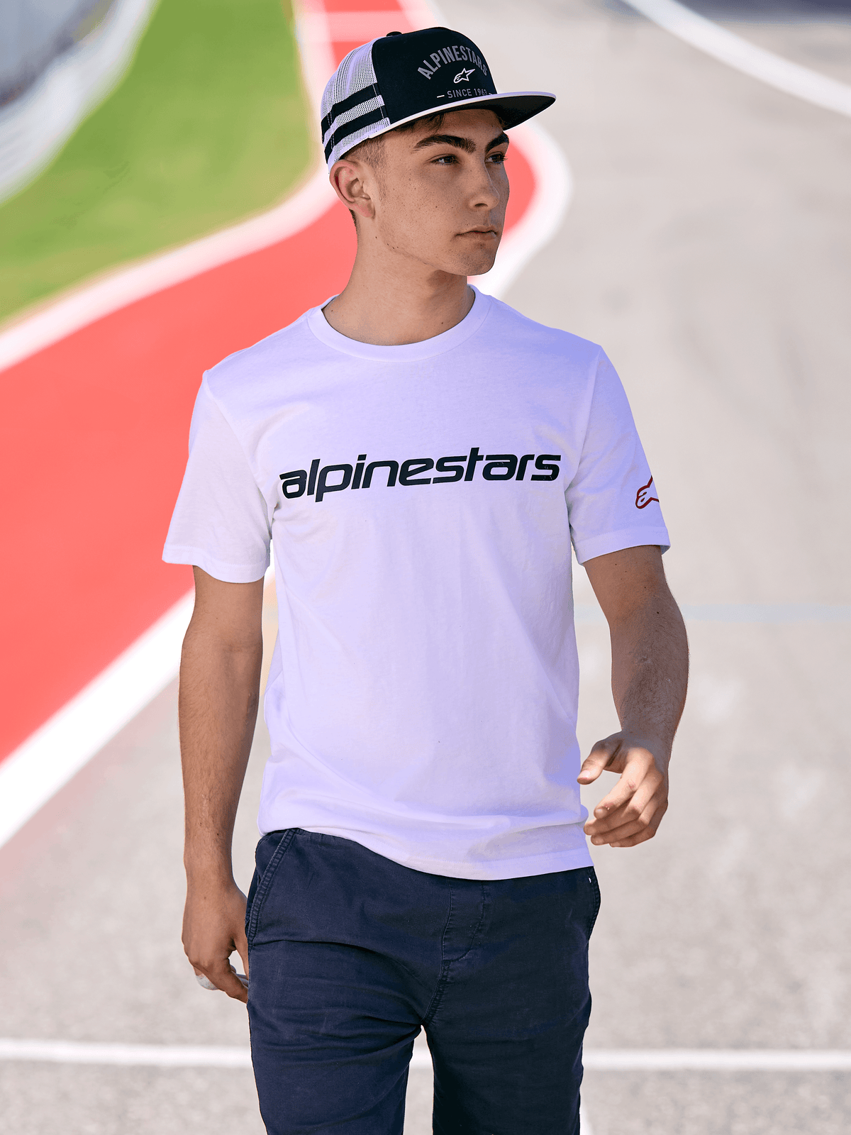 Uomo con cappello Alpinestars Backline Trucker in nero e bianco, abbinato a una t-shirt bianca con logo Alpinestars e pantaloni blu scuro, stile streetwear casual motorsport su sfondo pista all'aperto