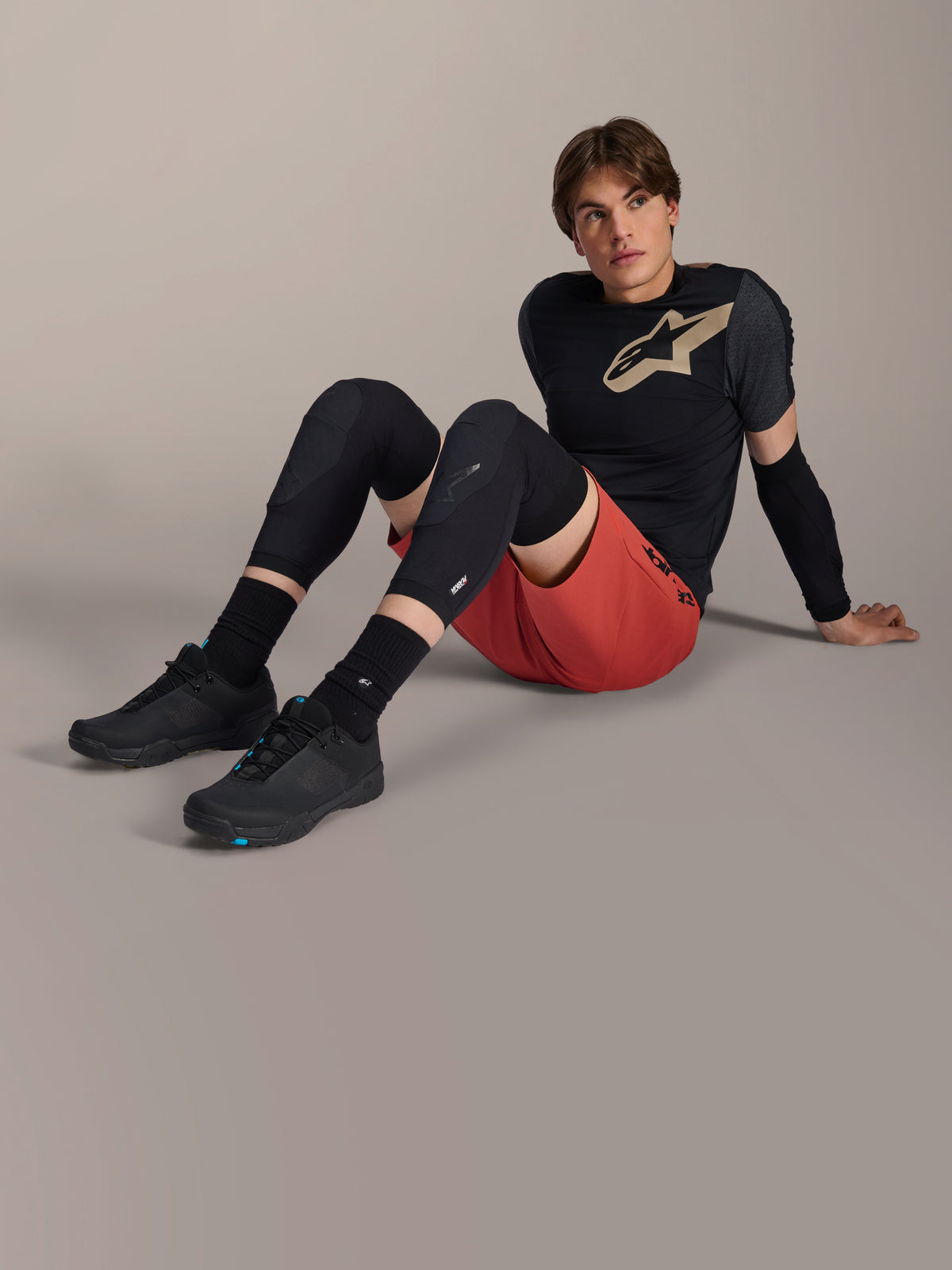 Gomitiera Alpinestars A-Motion Plasma, indossata da un ciclista seduto sul pavimento di uno studio, caratterizzata da una struttura in rete nera, abbinata a pantaloncini rossi e una maglia Alpinestars nera con logo beige, protezione per mountain bike ad alte prestazioni per trail riding.