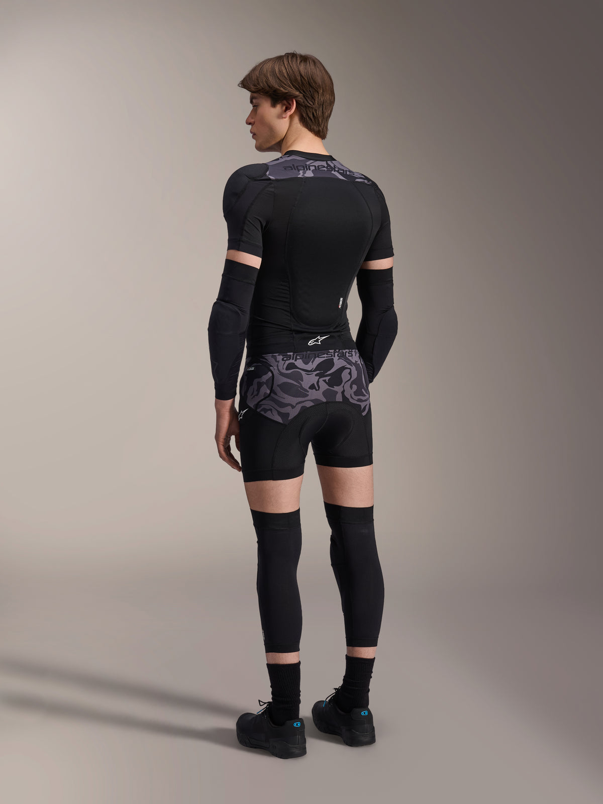 Modello che indossa i boxer protettivi Alpinestars A-Motion Plasma in Black Camo e la giacca protettiva A-Motion Plasma coordinata, vista posteriore con motivo mimetico grigio, gomitiere, ginocchiere e scarpe da ciclismo, set completo di protezioni per mountain bike