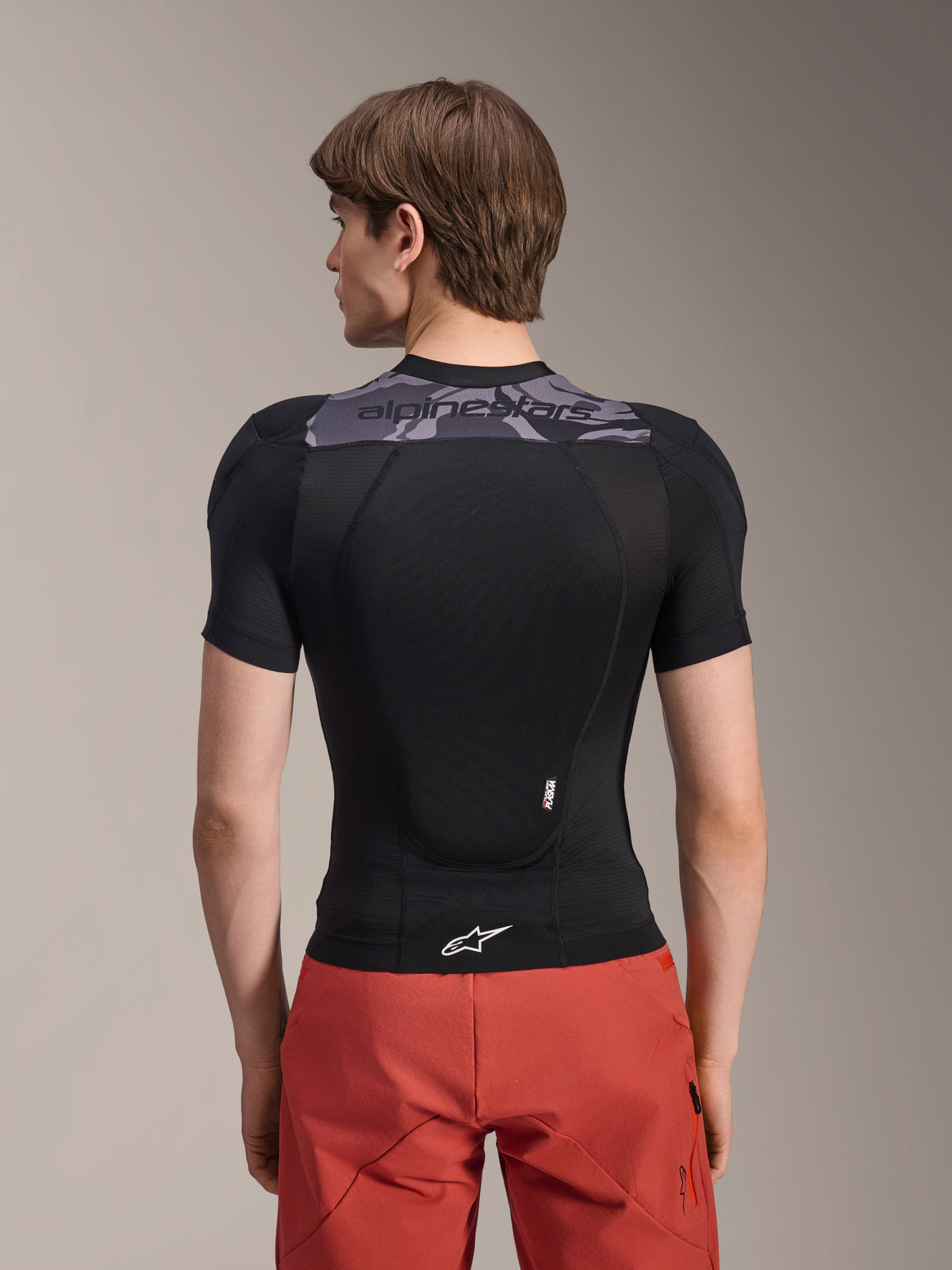 Giacca protettiva Alpinestars A-Motion Plasma - Maniche Corte, nero con dettagli mimetici grigi sulla parte superiore della schiena, vista posteriore con paraschiena integrato e logo Astars bianco sull'orlo, indossata con pantaloncini rossi, abbigliamento protettivo tecnico per mountain bike