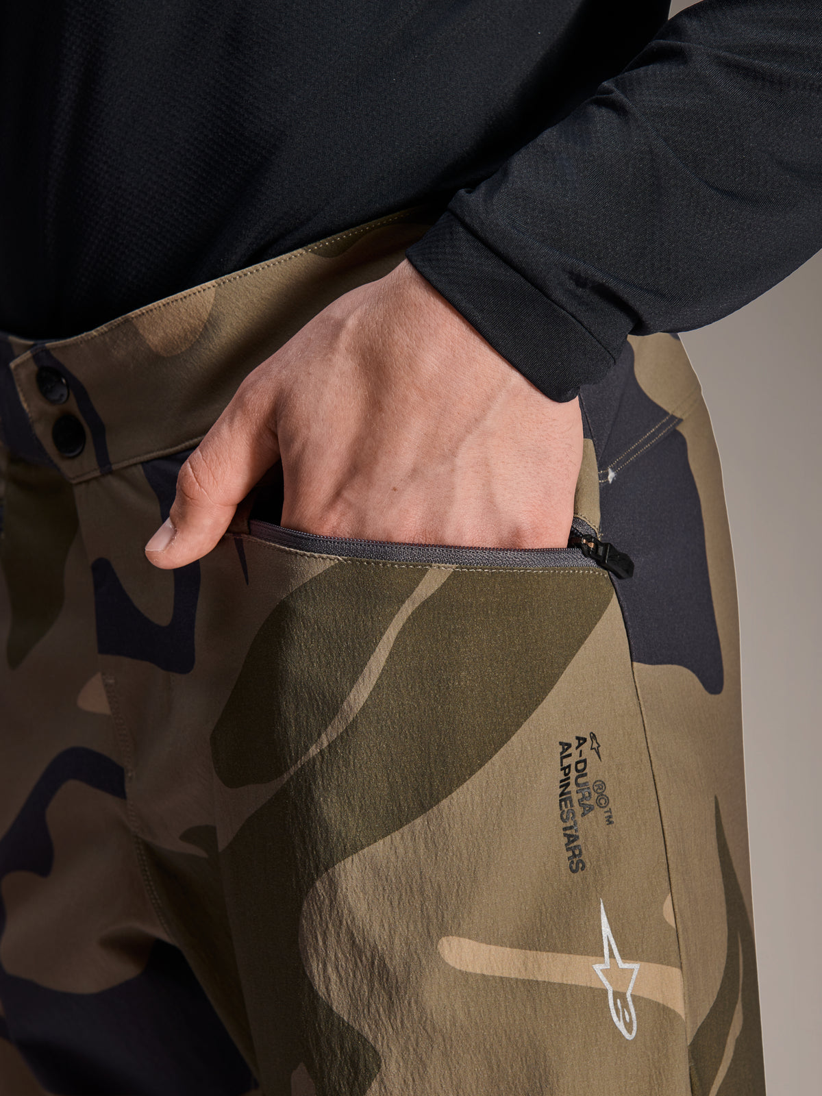 Dettaglio ravvicinato dei pantaloni Alpinestars A-Dura Camo, pantaloni da mountain bike mimetici marroni, con una mano in una tasca con cerniera, doppia chiusura a scatto in vita e logo Alpinestars sulla coscia