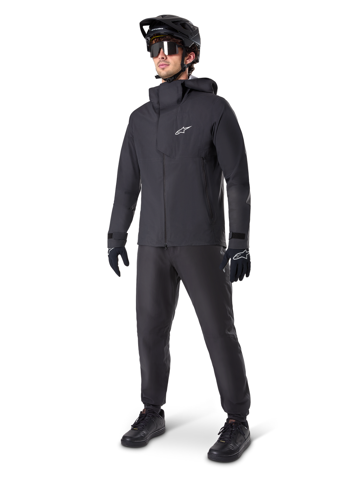 Mountain biker uomo con pantaloni impermeabili Alpinestars A-Dura neri, abbinati a giacca impermeabile nera con cappuccio coordinata, casco da MTB nero con visiera, occhiali da ciclismo scuri e guanti neri, completo professionale per trail riding in ogni condizione