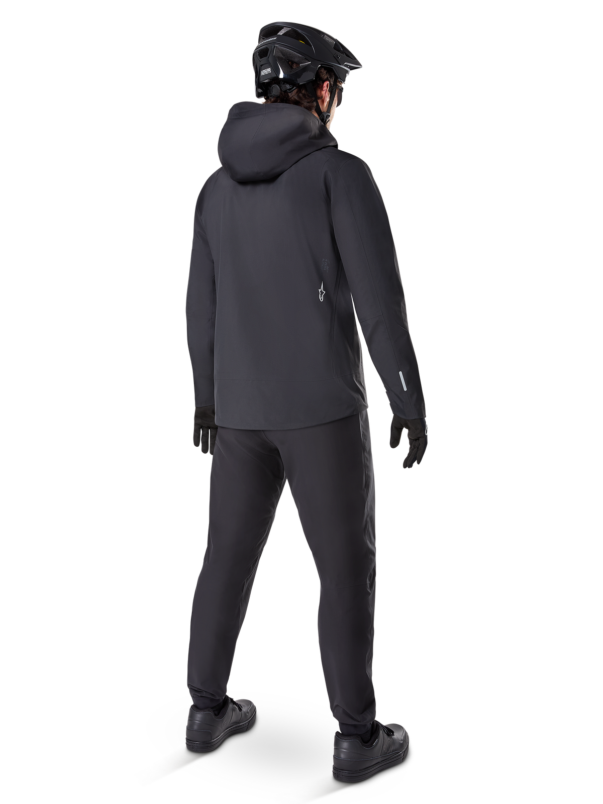 Pantaloni impermeabili Alpinestars A-Dura, pantaloni da MTB, nero, visti dal retro su un ciclista con abbigliamento da mountain bike nero completo di giacca con cappuccio e casco, abbigliamento impermeabile resistente per sentieri fangosi