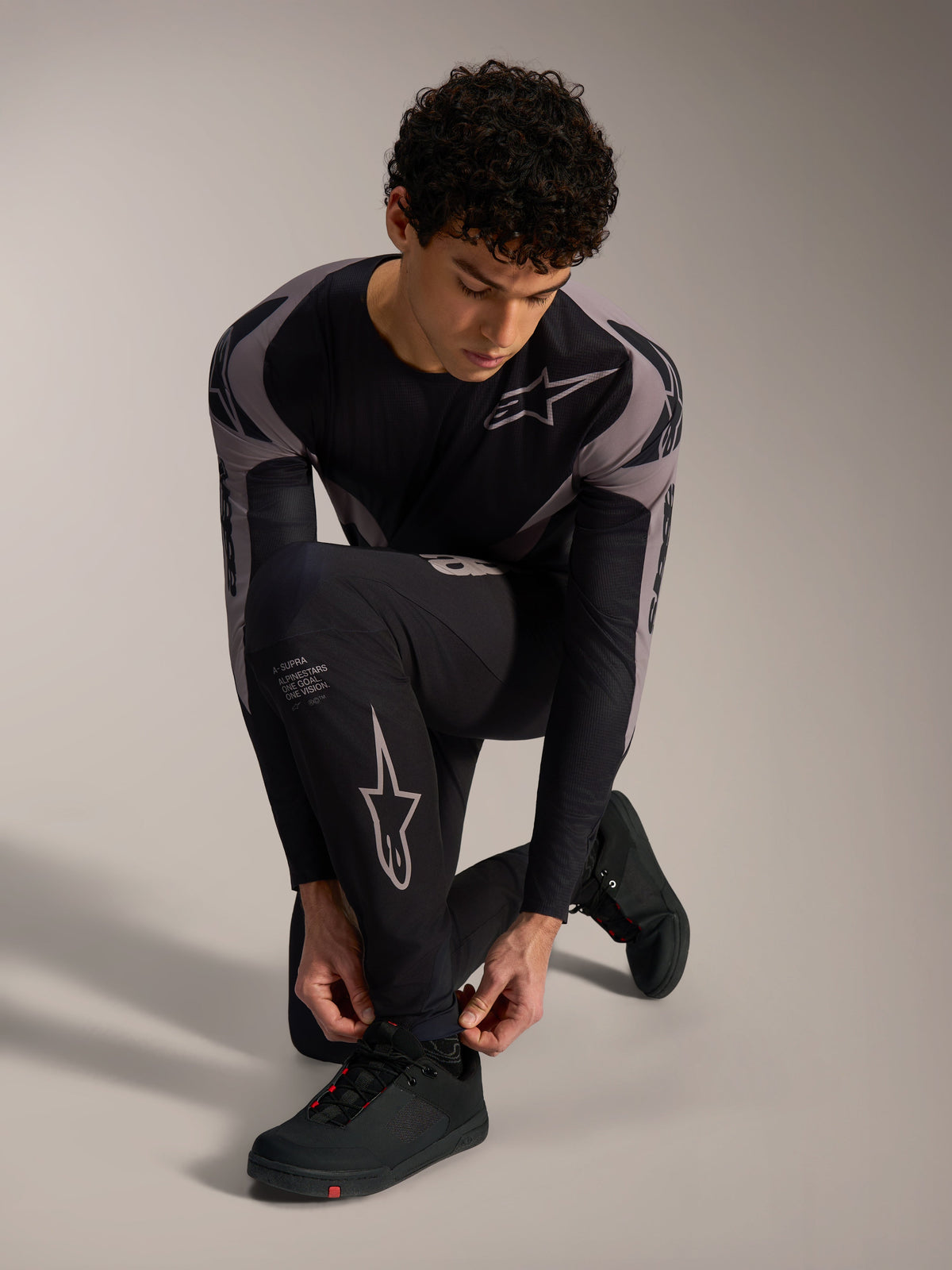 Pantaloni Alpinestars A-Supra Elite neri con loghi racing grigi, indossati da un modello inginocchiato per MTB.