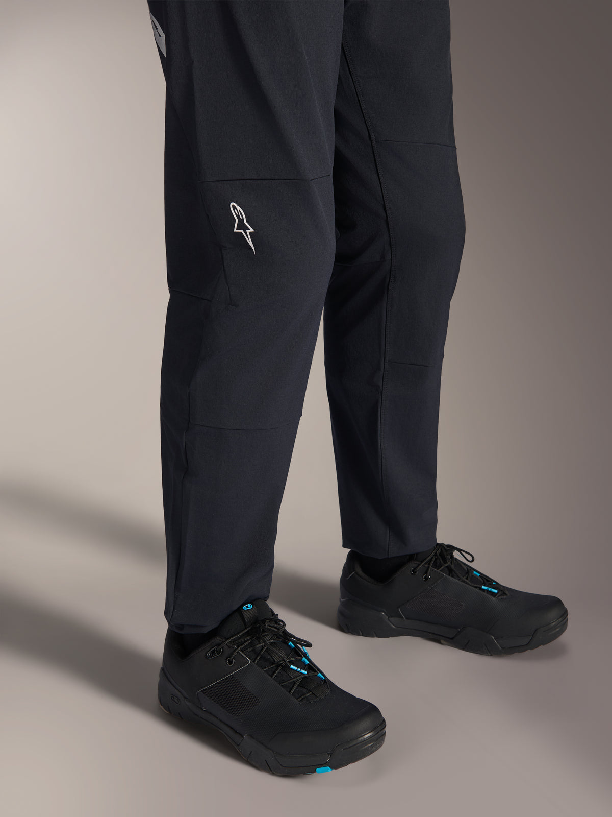 Pantaloni Alpinestars A-Dura Elite, pantaloni da mountain bike neri, vista della parte inferiore della gamba con logo Alpinestars bianco sul ginocchio, indossati con scarpe da ciclismo nere e blu, costruzione resistente per guida MTB