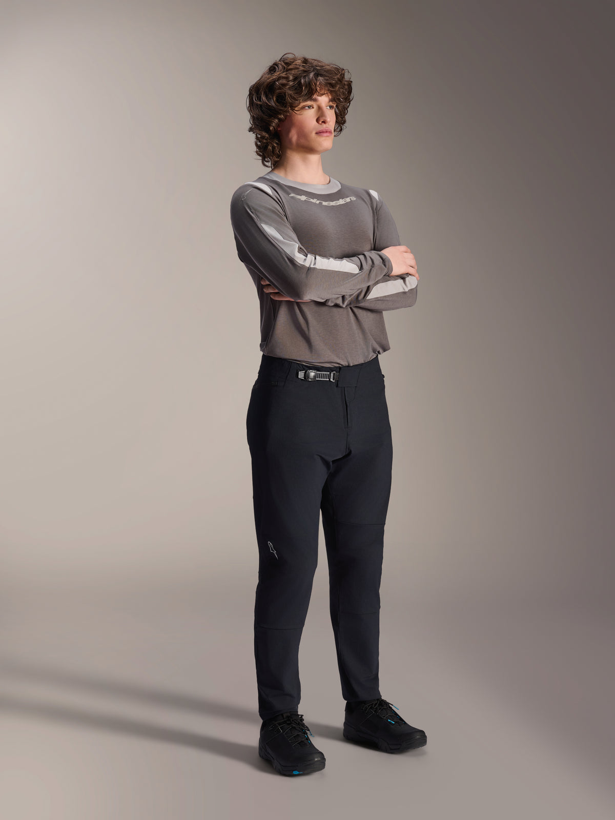 Modello uomo con pantaloni Alpinestars A-Dura Elite, pantaloni MTB neri, abbinati a una maglia grigia a maniche lunghe e scarpe da ciclismo nere, sistema di chiusura anteriore a cricchetto, abbigliamento professionale da mountain bike per trail ed enduro