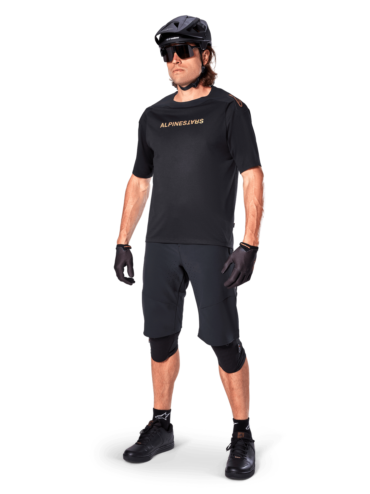 Ciclista con pantaloncini Alpinestars A-Aria Elite, pantaloncini da mountain bike neri, abbinati a una maglia nera con logo dorato, casco nero, occhiali da sole e ginocchiere, outfit tecnico completo per ciclismo fuoristrada