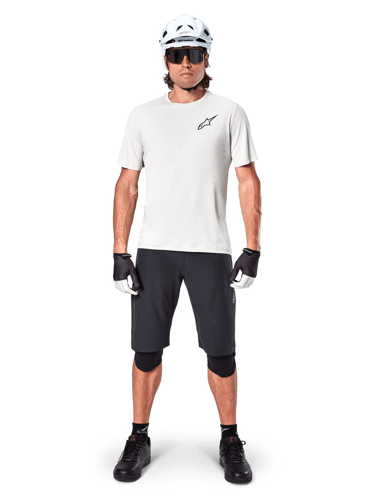 Mountain biker uomo con pantaloncini Alpinestars A-Aria neri, maglia tecnica bianca, casco bianco e protezioni per le ginocchia nere, equipaggiamento professionale da ciclismo fuoristrada in vista frontale a figura intera