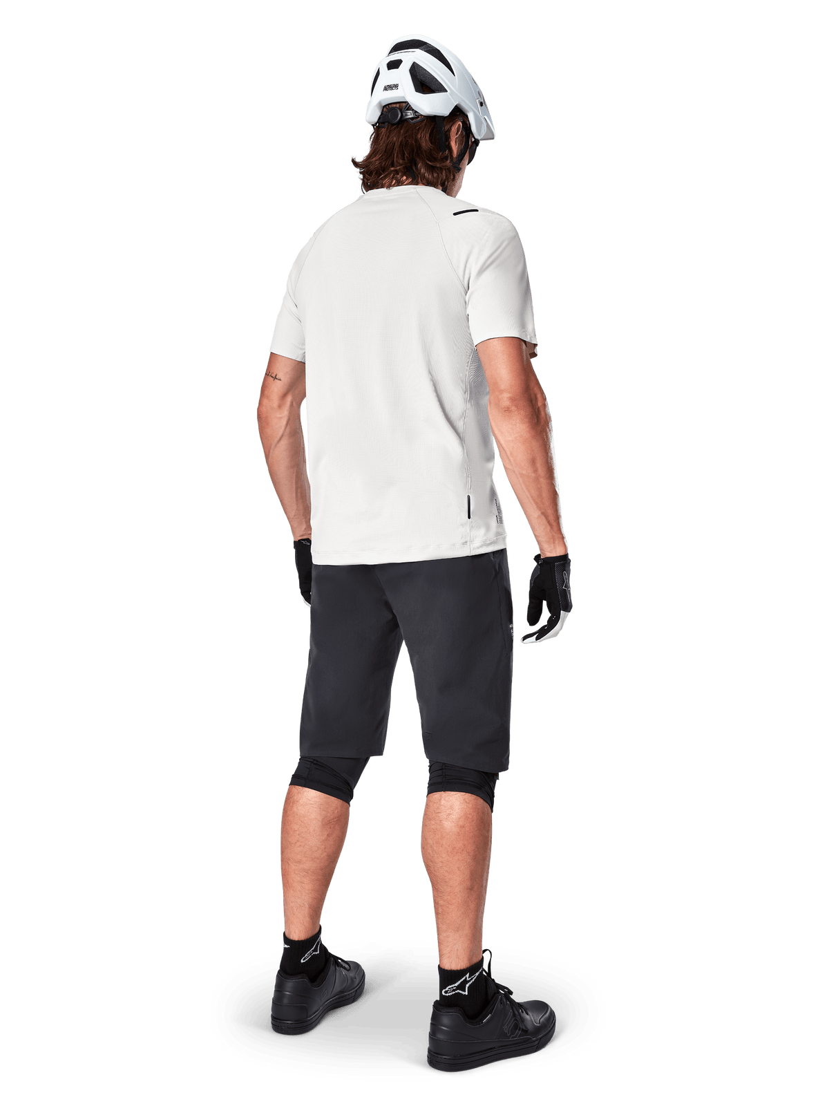Mountain biker visto da dietro con pantaloncini Alpinestars A-Aria in nero, maglia tecnica bianca a maniche corte, casco bianco, guanti neri e scarpe da mountain bike nere, abbigliamento da ciclismo per trail riding