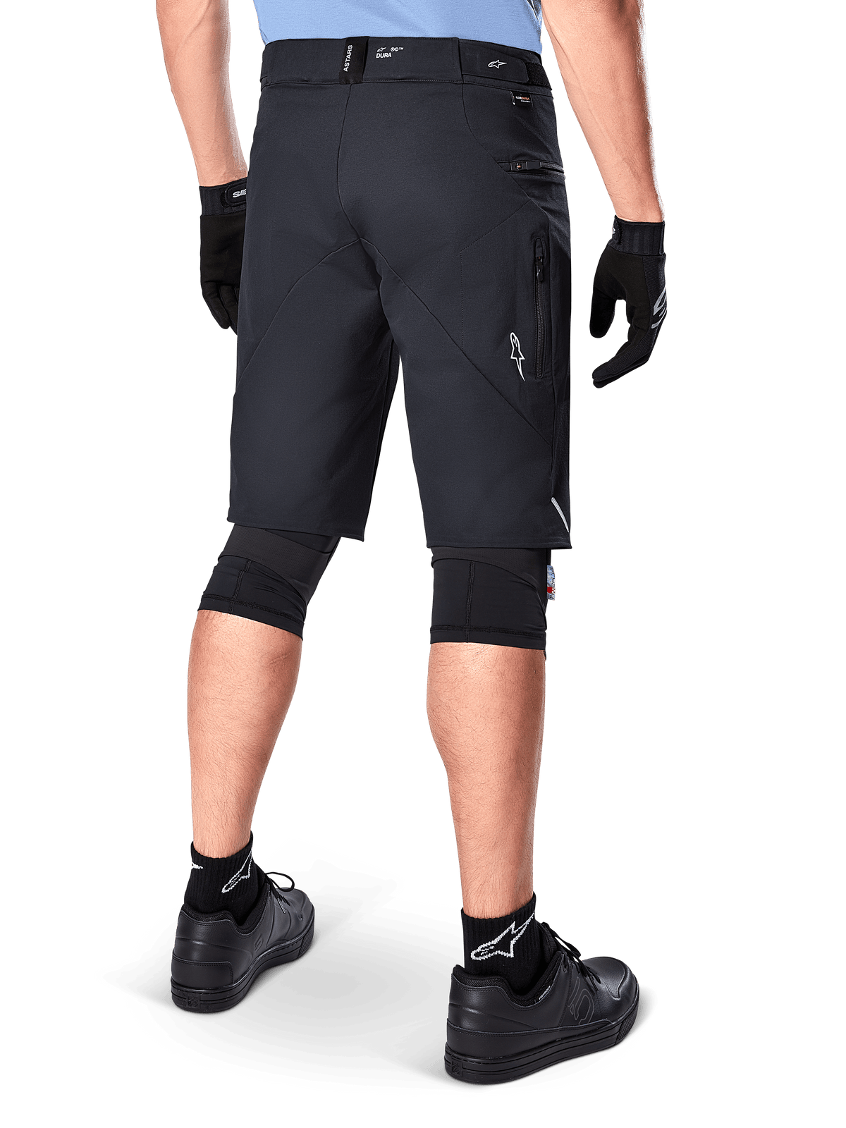 Pantaloncini Alpinestars A-Dura Elite, nero, vista posteriore di un ciclista con abbigliamento da mountain bike, incluse ginocchiere nere, guanti e calze con loghi bianchi, con tasche laterali con zip e accenti riflettenti in resistente tessuto Cordura.