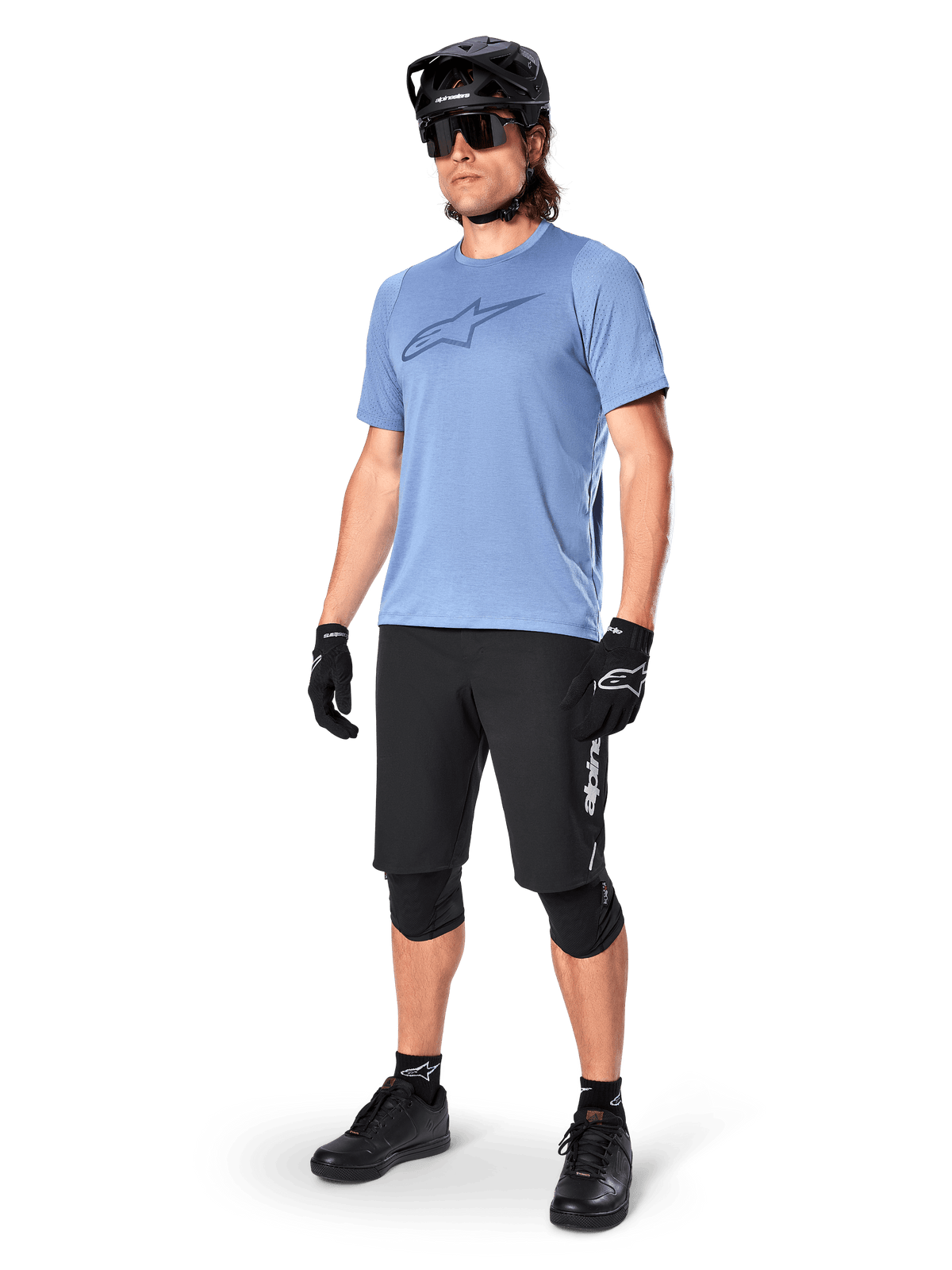 Ciclista con pantaloncini Alpinestars A-Dura Elite neri, maglia a maniche corte azzurra con logo sul petto, casco nero, occhiali da sole e ginocchiere protettive, equipaggiamento completo da mountain bike per percorsi fuoristrada.