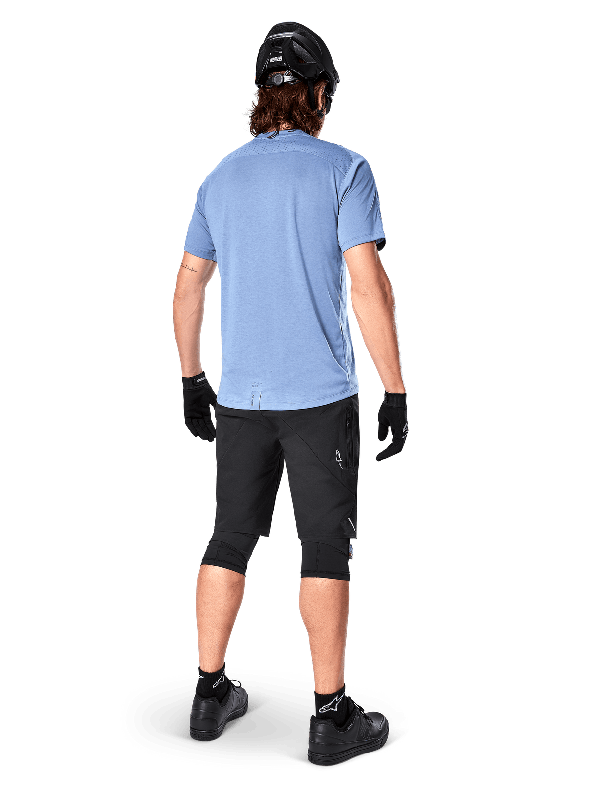 Alpinestars A-Dura Elite Shorts, pantaloncini da mountain bike, nero, vista posteriore su modello, indossati con maglia azzurra a maniche corte e attrezzatura protettiva nera, design tecnico con tasche con zip e dettagli riflettenti per il ciclismo fuoristrada