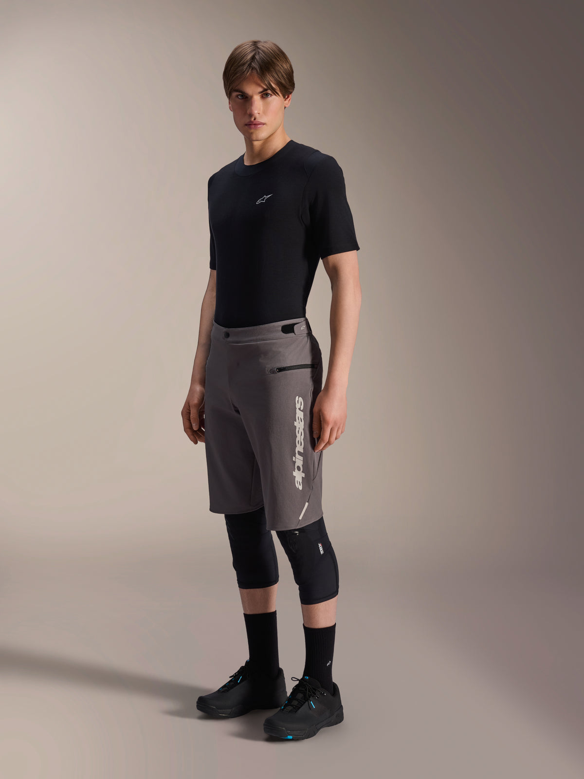 Alpinestars A-Dura Elite Shorts, pantaloncini da mountain bike grigio scuro, indossati da un modello con t-shirt nera e ginocchiere, con logo Alpinestars bianco verticale sulla gamba sinistra, tessuto tecnico Cordura per ciclismo fuoristrada