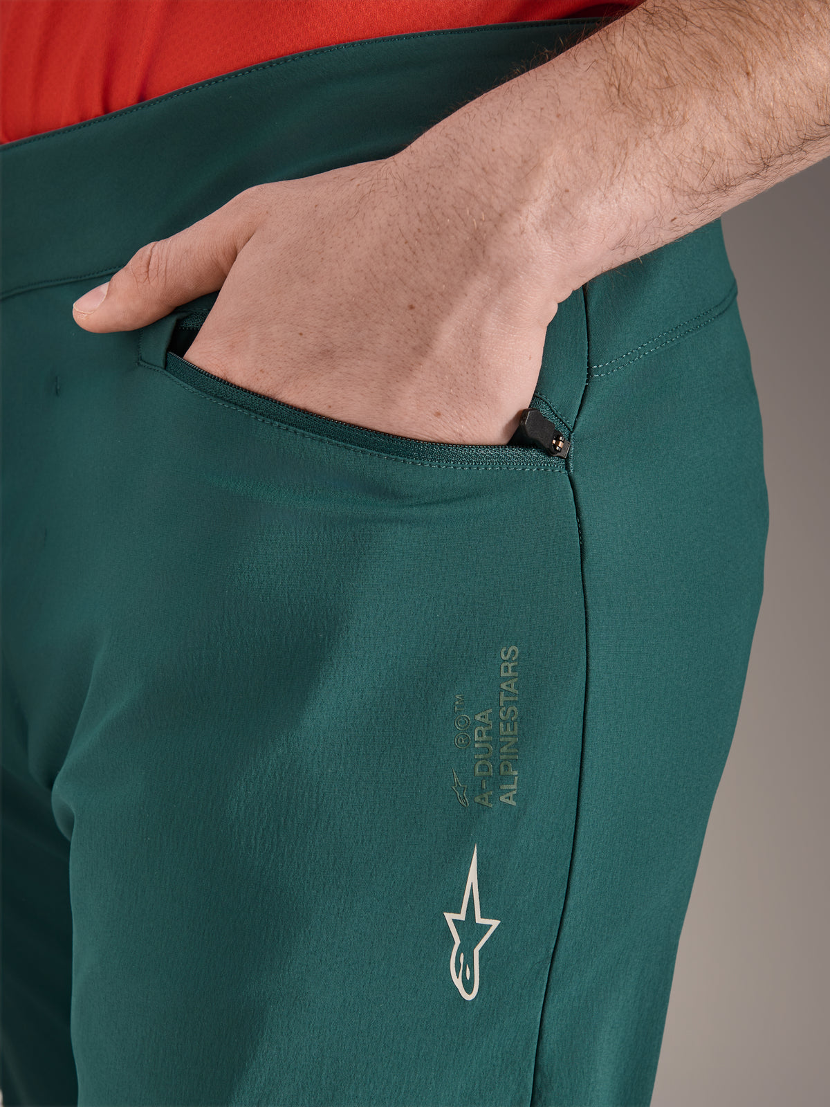 Pantaloni Alpinestars A-Dura in verde Dark Pine, primo piano della tasca con cerniera e mano, con logo Alpinestars bianco e scritta sulla coscia, pantaloni da ciclismo MTB resistenti