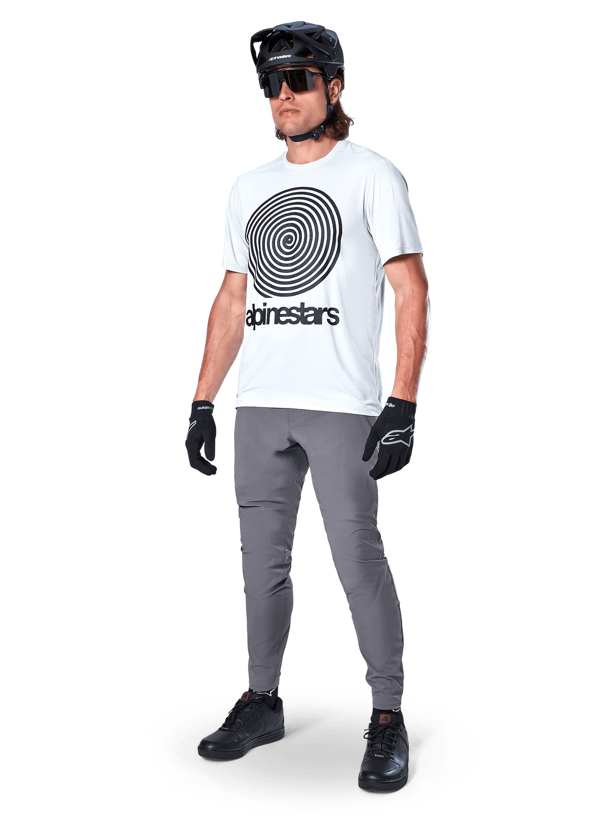 Pantaloni Alpinestars A-Dura, pantaloni da MTB, grigio scuro, indossati con t-shirt bianca con grafica a spirale, casco da MTB nero, occhiali da sole e guanti neri, tessuto resistente elasticizzato in 2 direzioni per il ciclismo fuoristrada