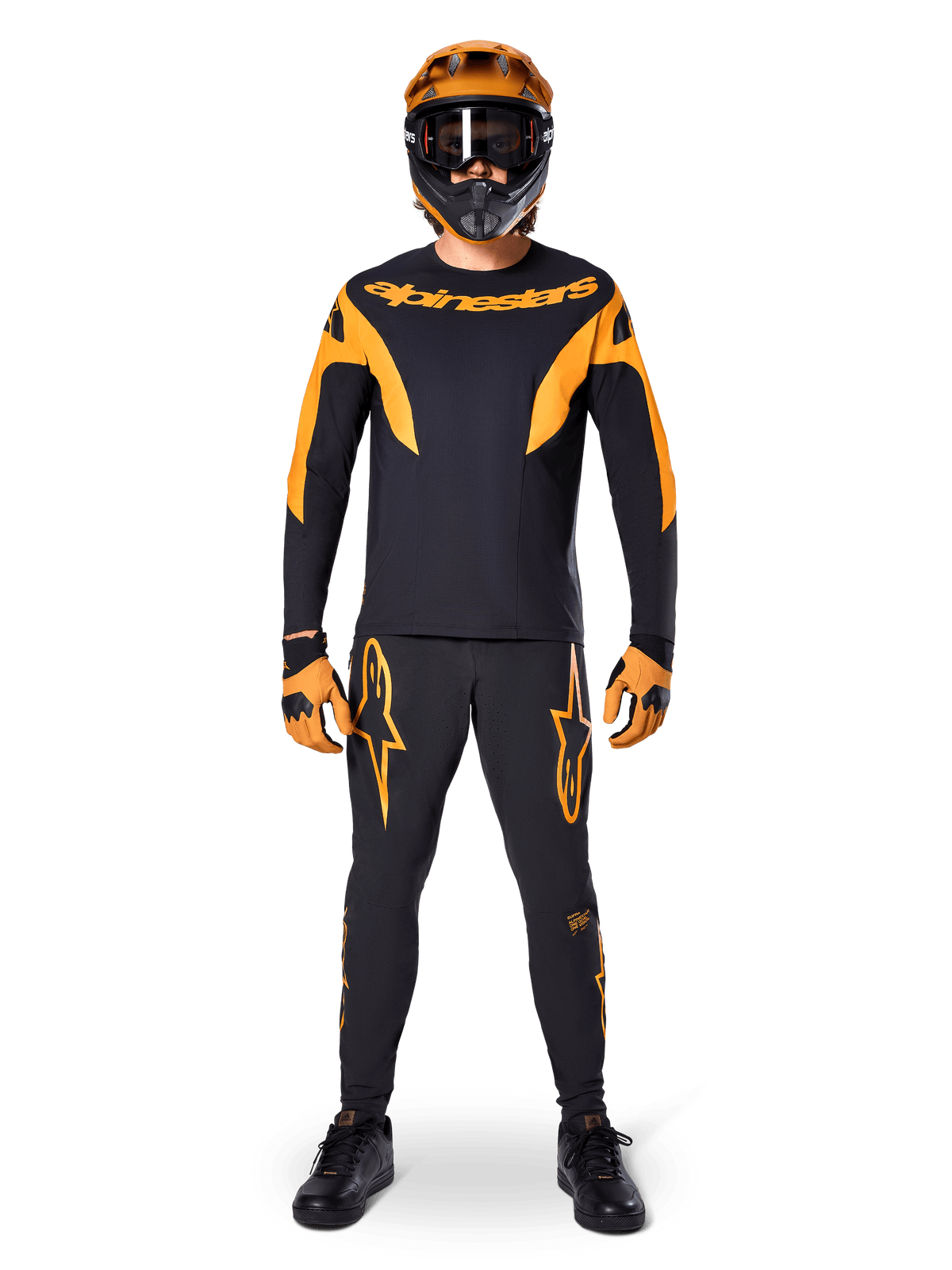 Pantaloni Alpinestars A-Supra Race, pantaloni da gara MTB, nero e arancione, indossati da un pilota con un kit da gara completo composto da maglia nera e arancione, casco arancione e guanti coordinati, con grandi loghi a stella arancioni sulle gambe