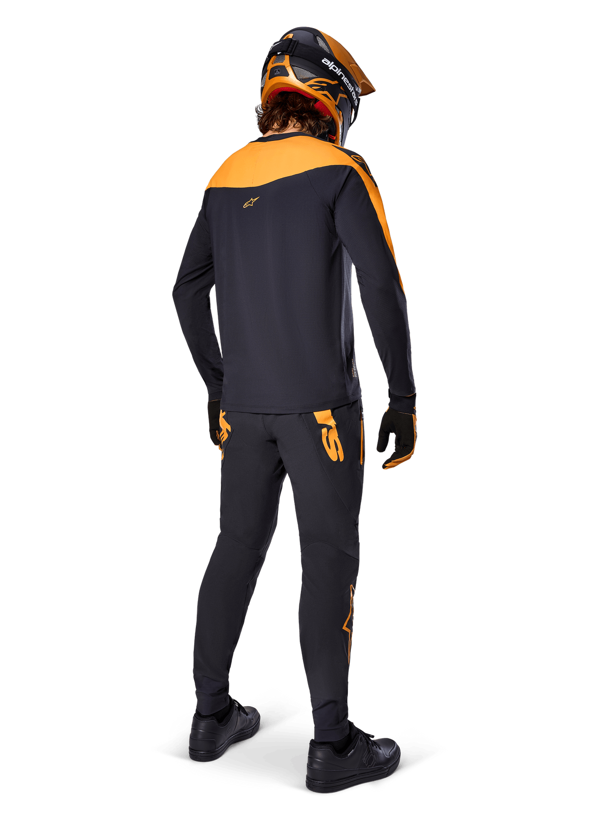 Alpinestars A-Supra Race Pants, pantaloni da gara MTB, neri con loghi arancioni, vista posteriore su un rider che indossa maglia, casco e guanti coordinati nero e arancione, evidenziando il wordmark ispirato al MX sul carré posteriore e la vestibilità performante per trail tecnici.