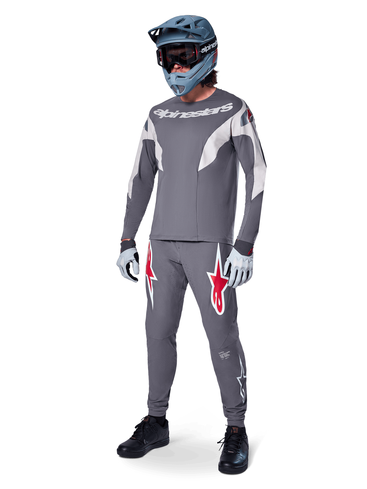 Alpinestars pantaloni A-Supra Race in grigio scuro, con vistosi loghi rossi e bianchi sulle cosce, indossati da un rider come parte di un kit completo da mountain bike con maglia grigia coordinata, casco integrale grigio-blu e guanti grigio chiaro, abbigliamento ad alte prestazioni per gare MTB