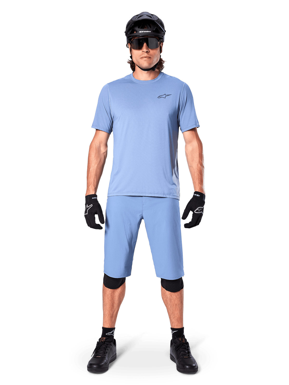 Modello con pantaloncini Alpinestars A-Dura in Infinity Blue, maglia azzurra coordinata, casco da mountain bike nero, occhiali da sole neri, guanti e ginocchiere neri, outfit completo da MTB per trail riding