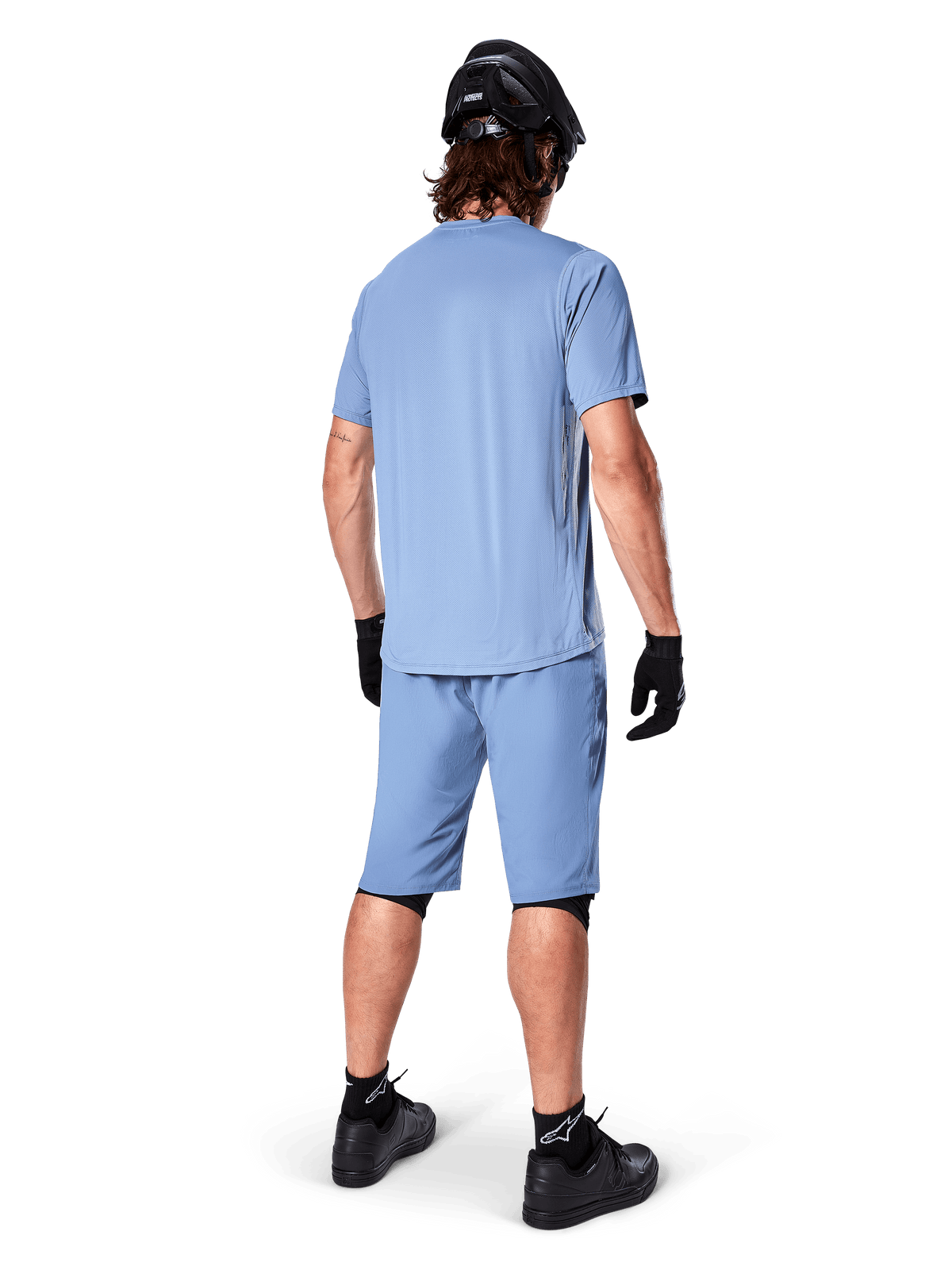 Vista posteriore di un mountain biker con pantaloncini Alpinestars A-Dura in Infinity Blue, maglia a maniche corte coordinata, casco, guanti e scarpe neri, abbigliamento tecnico coordinato per trail riding in MTB