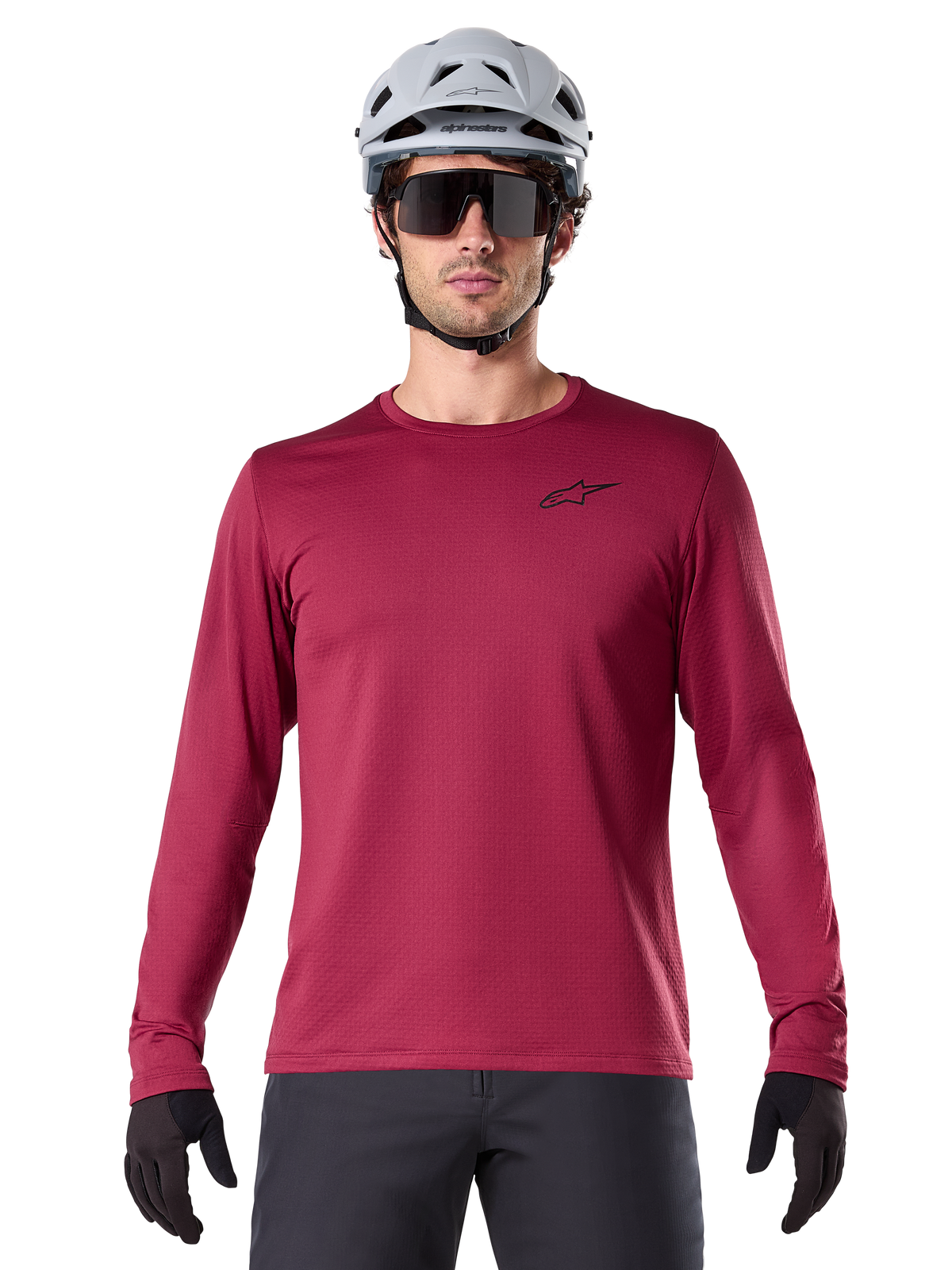 Ciclista uomo che indossa la maglia termica Alpinestars A-Dura, maglia MTB a maniche lunghe bordeaux, abbinata a un casco grigio e occhiali da sole neri, piccolo logo a stella nero sul petto, tessuto in pile isolante per mountain bike in climi freddi