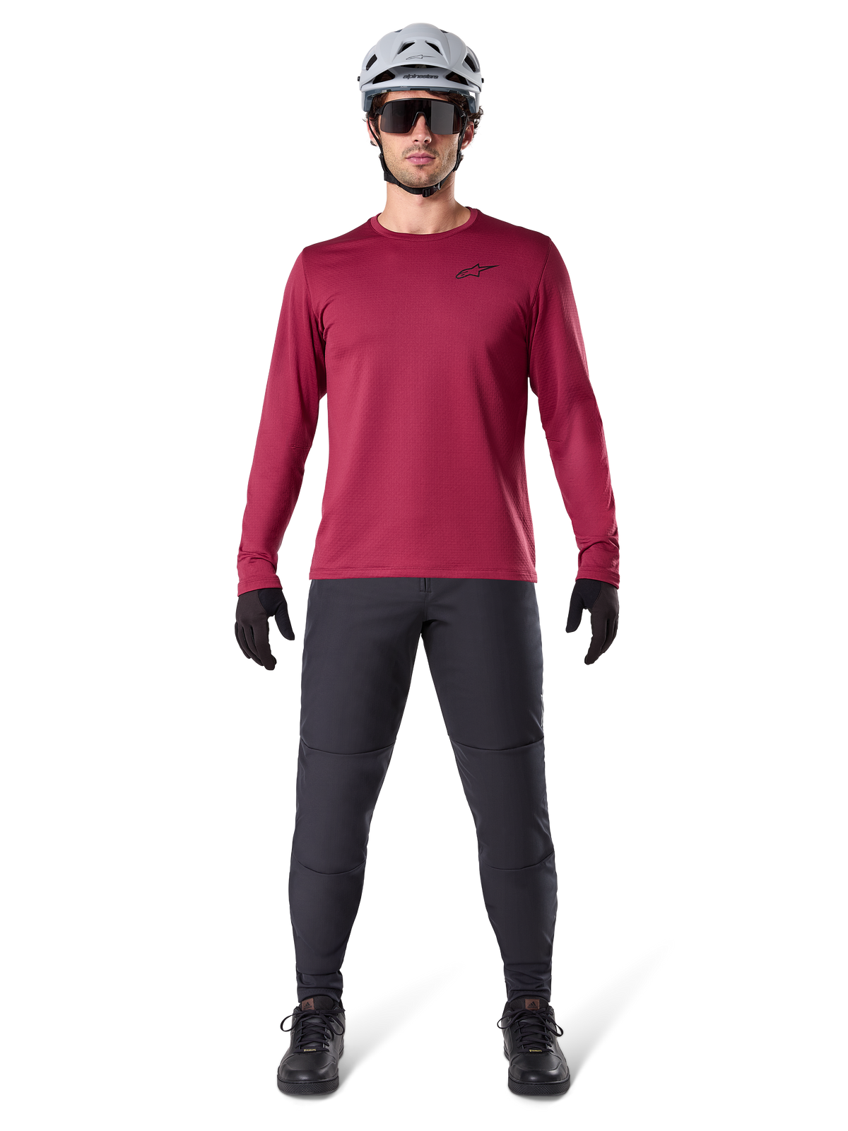 Modello uomo che indossa la maglia termica Alpinestars A-Dura colore Burgundy, maglia da ciclismo isolante a maniche lunghe, abbinata a pantaloni da mountain bike grigio scuro, casco bianco, occhiali da sole neri e guanti neri, equipaggiamento professionale da MTB per il freddo