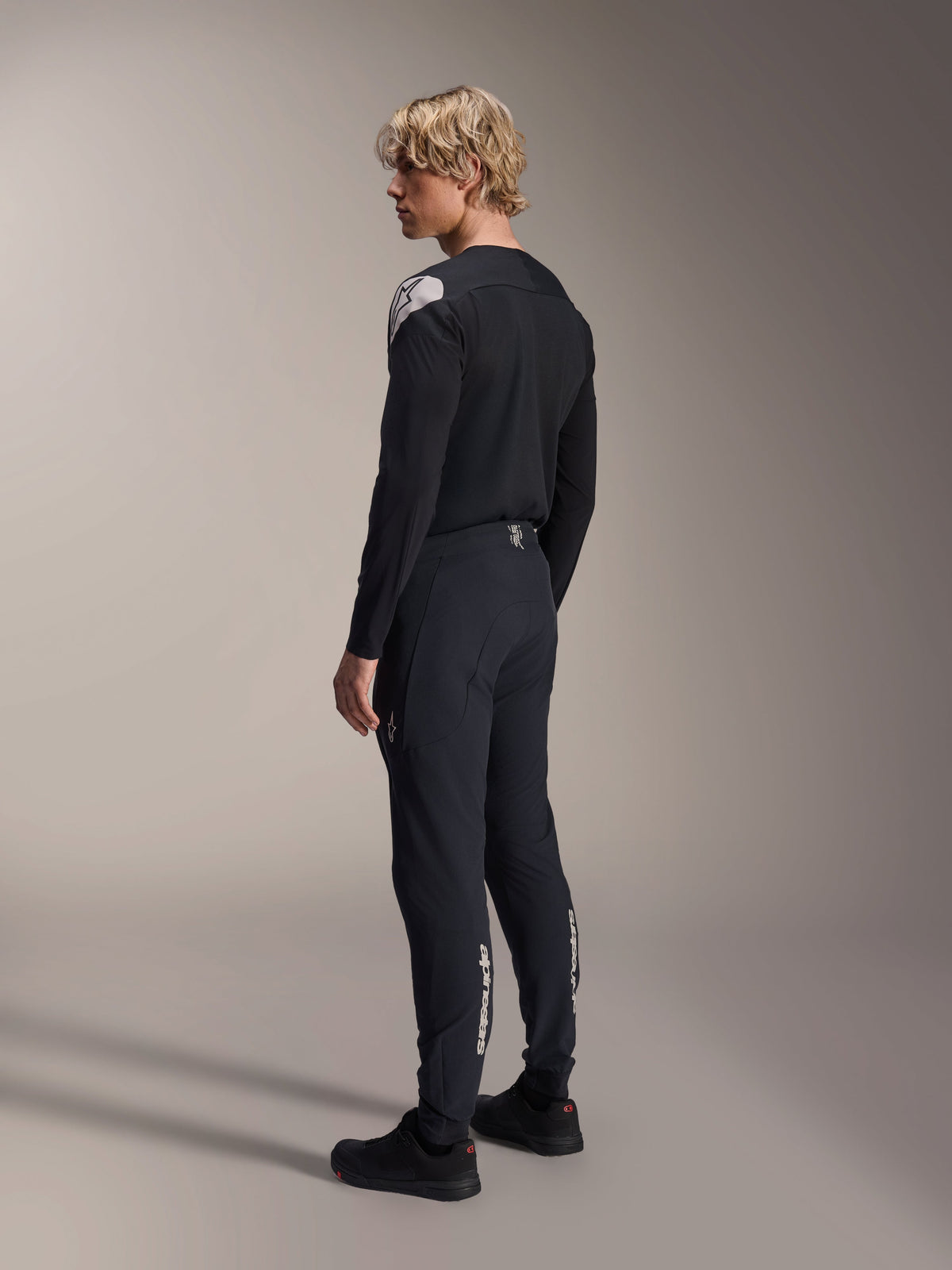 Maglia Alpinestars A-Aria Polartec Elite - Maniche Lunghe, nera, indossata da un modello in vista laterale posteriore, con logo bianco sulla spalla, abbinata a pantaloni da ciclismo neri coordinati per un completo professionale da MTB.