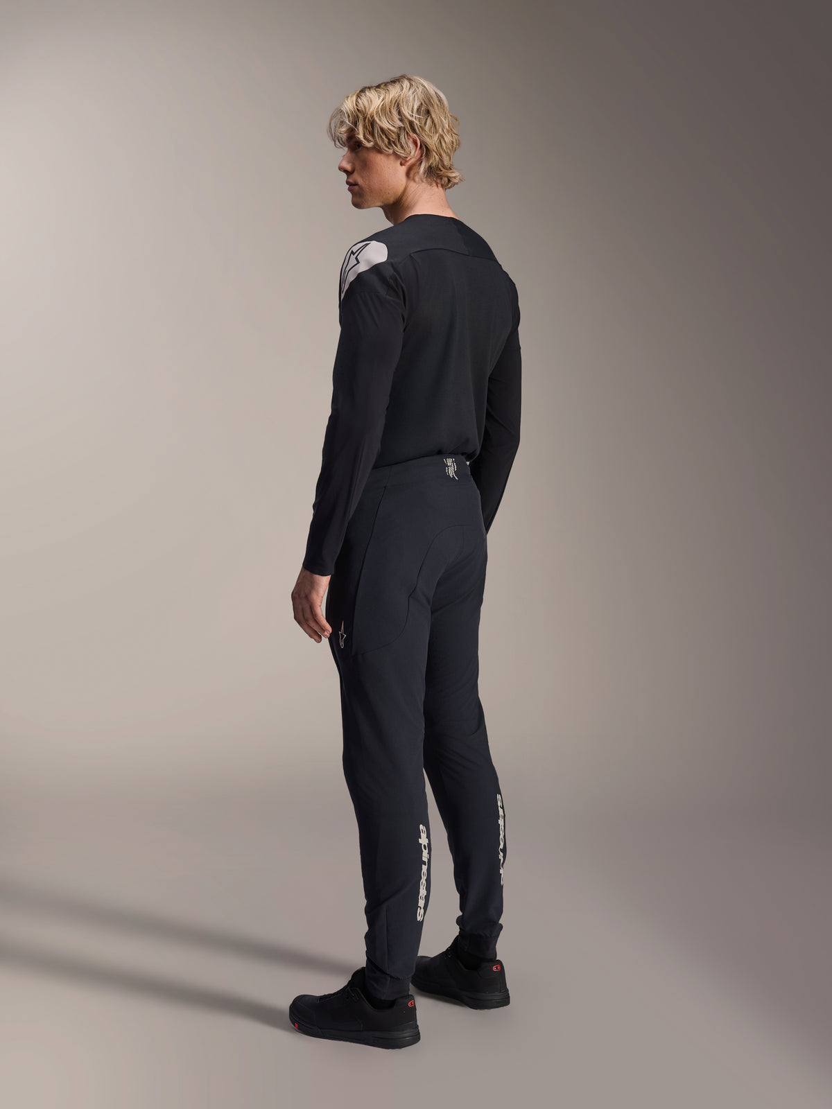 Pantaloni Alpinestars A-Aria Elite, pantaloni da mountain bike, Black Titanium, vista posteriore a tre quarti su modello, caratterizzati dal marchio Alpinestars bianco sulla parte inferiore delle gambe, vestibilità performante con polsini elastici, abbinati a una maglia coordinata nera a maniche lunghe.
