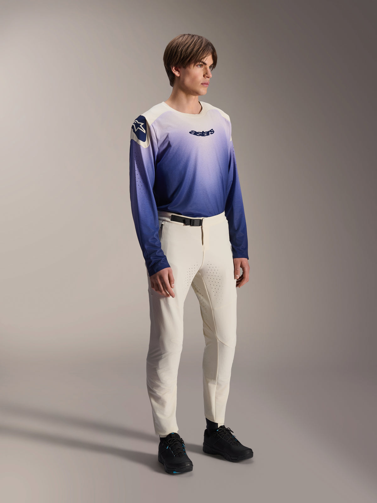 Modello che indossa i pantaloni Alpinestars A-Aria Elite in Ecru, pantaloni da mountain bike bianco sporco con ventilazione tagliata al laser e fibbia nera in vita, abbinati a una maglia a maniche lunghe con sfumatura blu e viola, abbigliamento tecnico da ciclismo per percorsi fuoristrada