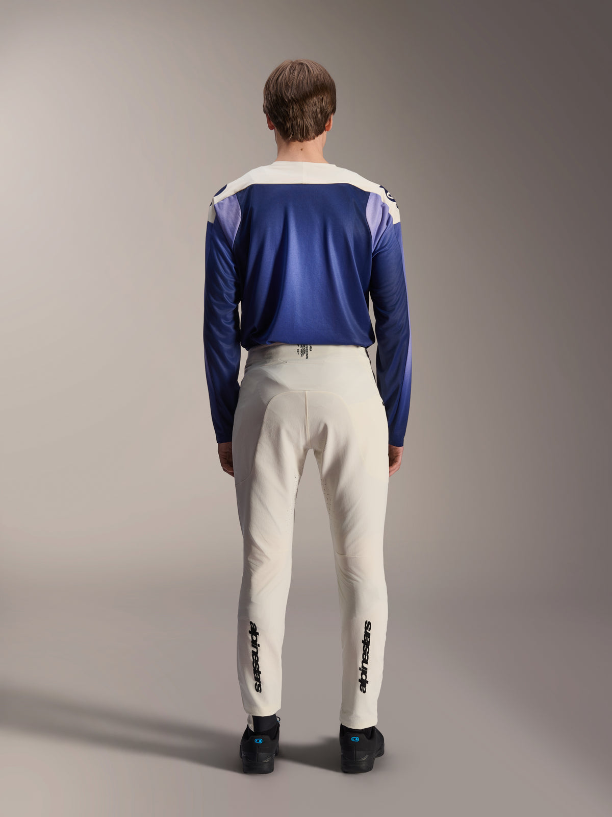 Alpinestars Pantaloni A-Aria Elite in Ecru, visti dal retro su un modello abbinati a una maglia a maniche lunghe A-Aria Polartec Maker Elite blu, pantaloni tecnici da MTB bianco panna con loghi neri sulla parte inferiore delle gambe, abbigliamento tecnico da ciclismo per mountain bike