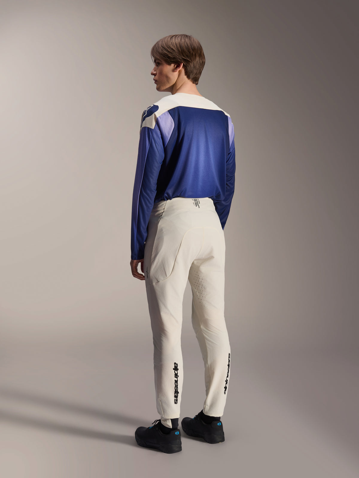 Pantaloni Alpinestars A-Aria Elite, pantaloni da MTB, color ecru, vista posteriore indossata da un modello, con marchio nero sulla parte inferiore delle gambe e perforazioni laser per la ventilazione, abbinati a una maglia a maniche lunghe blu e viola e scarpe nere, abbigliamento professionale da mountain bike