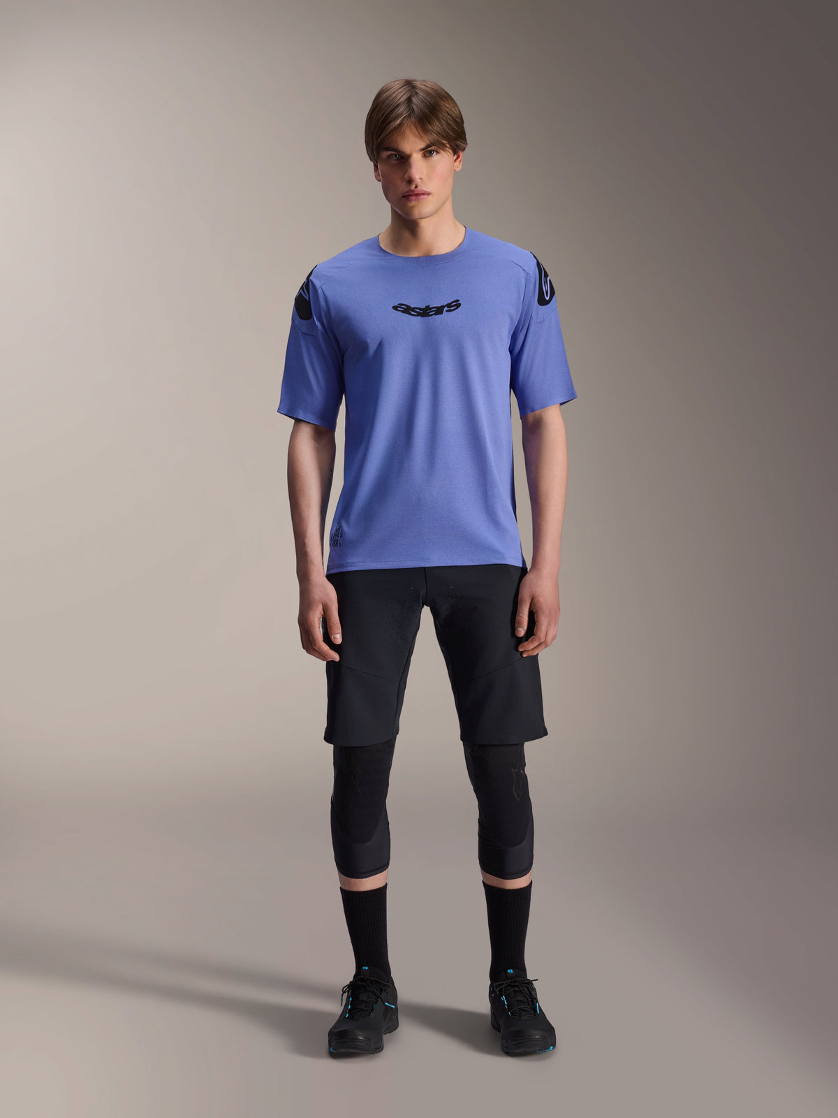 Modello maschile che indossa la maglia Alpinestars A-Aria Polartec Elite a maniche corte in Baja Blue, logo astars nero sul petto, abbinata a pantaloncini da MTB neri, ginocchiere e scarpe da ciclismo nere, scatto in studio per abbigliamento da mountain bike
