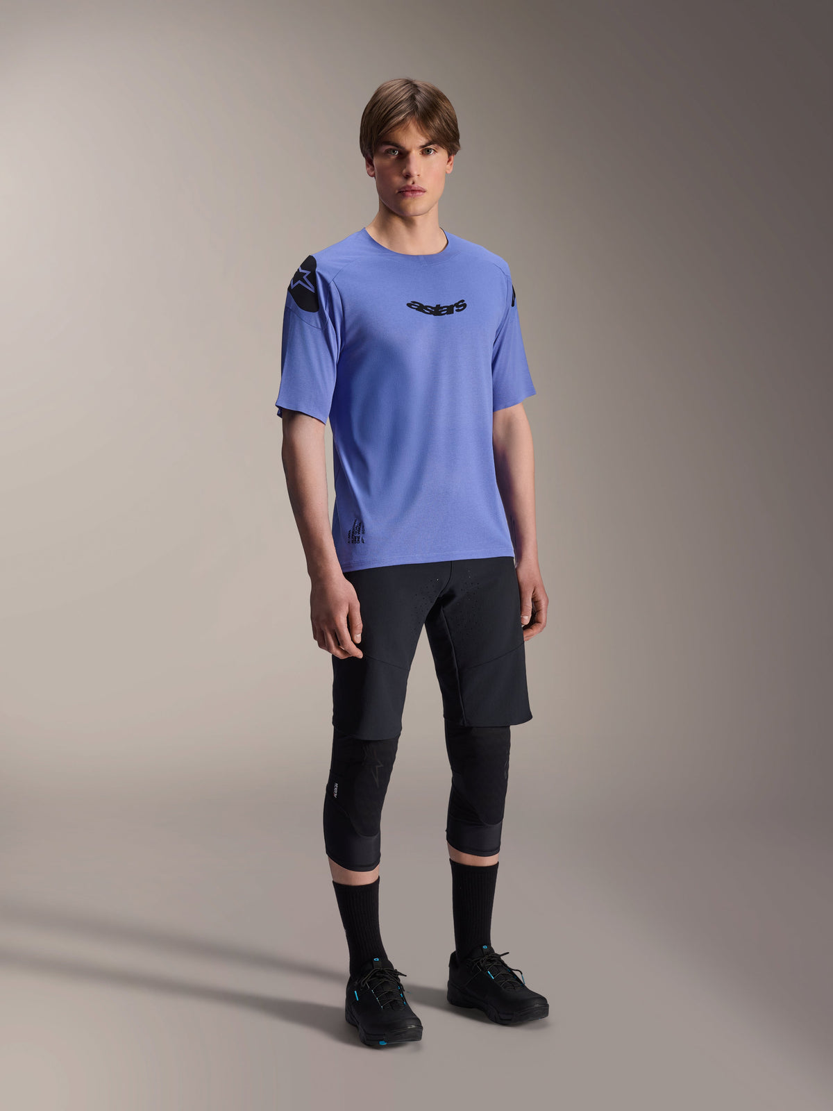 Modello che indossa la maglia a maniche corte Alpinestars A-Aria Polartec Elite in Baja Blue, abbinata a pantaloncini da MTB e ginocchiere neri, con loghi Astars neri sul petto e sulla spalla, abbigliamento tecnico da mountain bike ad alte prestazioni