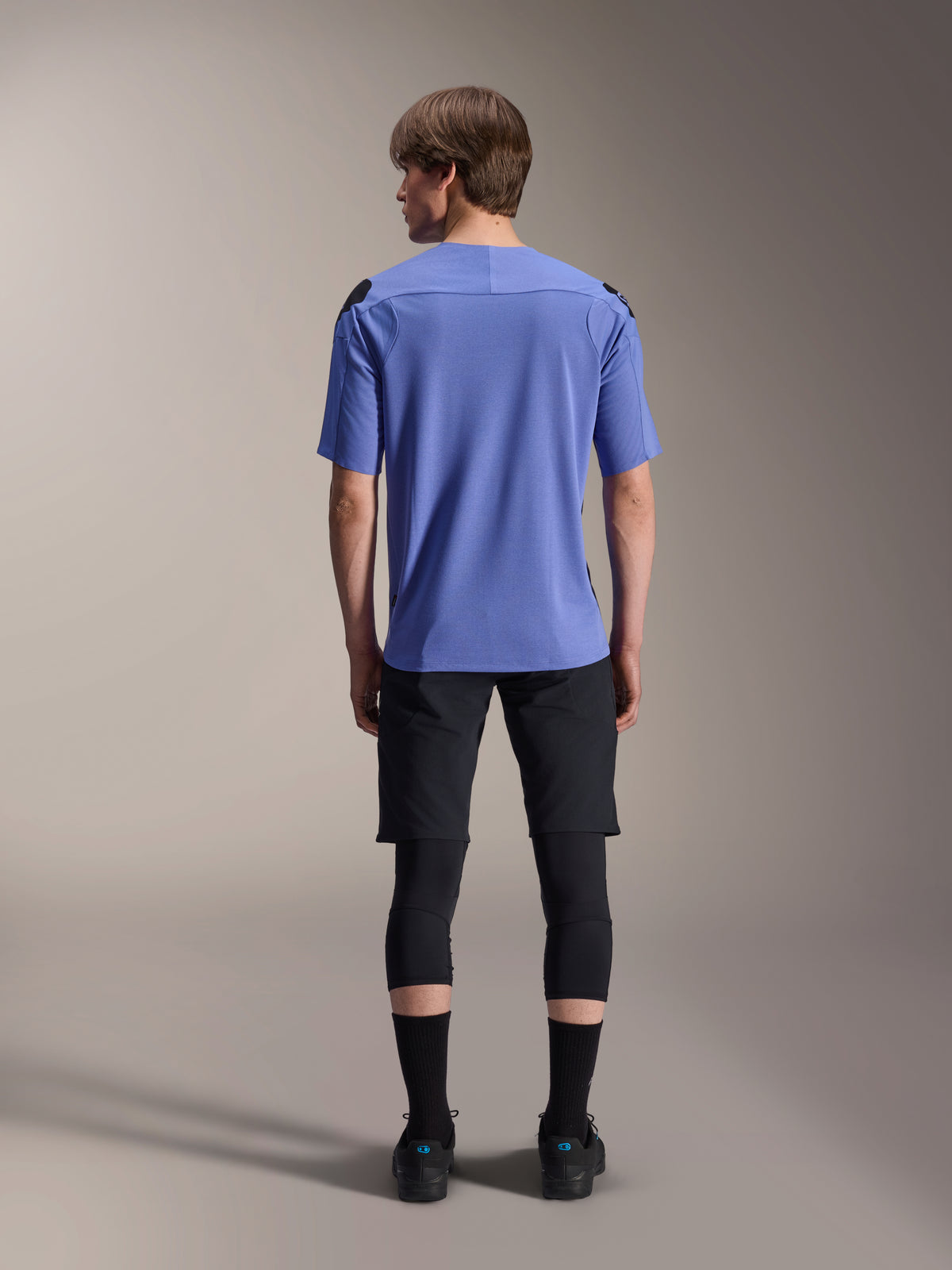 Ciclista di mountain bike visto da dietro che indossa pantaloncini Alpinestars A-Aria Lite neri, abbinati a una maglia blu a maniche corte, ginocchiere nere e calze nere, abbigliamento tecnico da ciclismo per percorsi fuoristrada