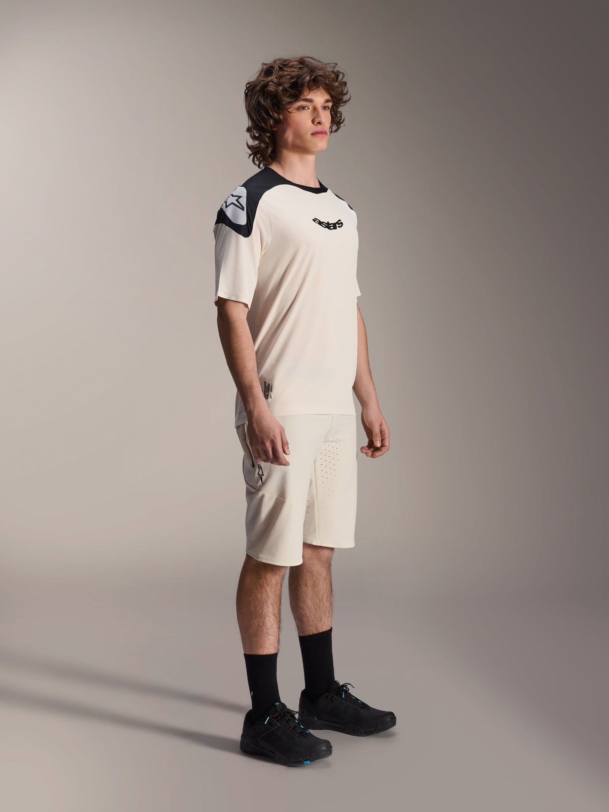 Alpinestars A-Aria Polartec Elite Short Sleeve Jersey, color ecru con pannelli sulle spalle e logo neri, indossata da un modello con pantaloncini coordinati e scarpe nere, abbigliamento tecnico da mountain bike per il trail riding fuoristrada.