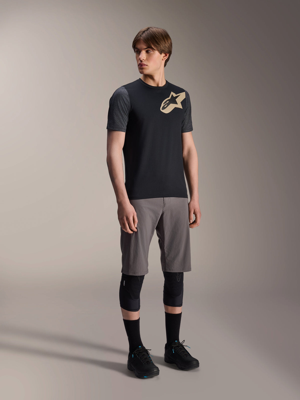 Modello uomo che indossa la maglia a maniche corte Alpinestars A-Dura Lipan nera, con maniche grigio scuro e logo Astars tan sul petto, abbinata a pantaloncini da mountain bike grigi, ginocchiere nere e scarpe da ciclismo nere, abbigliamento coordinato per mountain bike