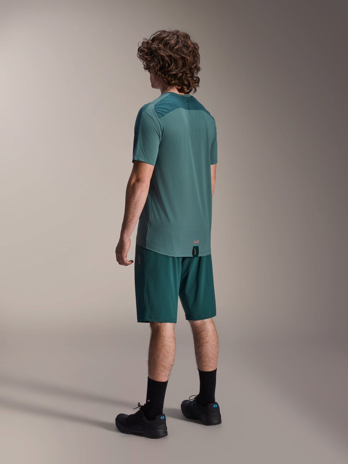 Modello che indossa la maglia a maniche corte Alpinestars A-Dura Lipan in Silver Pine, vista dal retro, abbinata a pantaloncini da mountain bike verde scuro coordinati, calze e scarpe nere, con pannello posteriore superiore testurizzato e orlo posteriore allungato per una maggiore copertura.