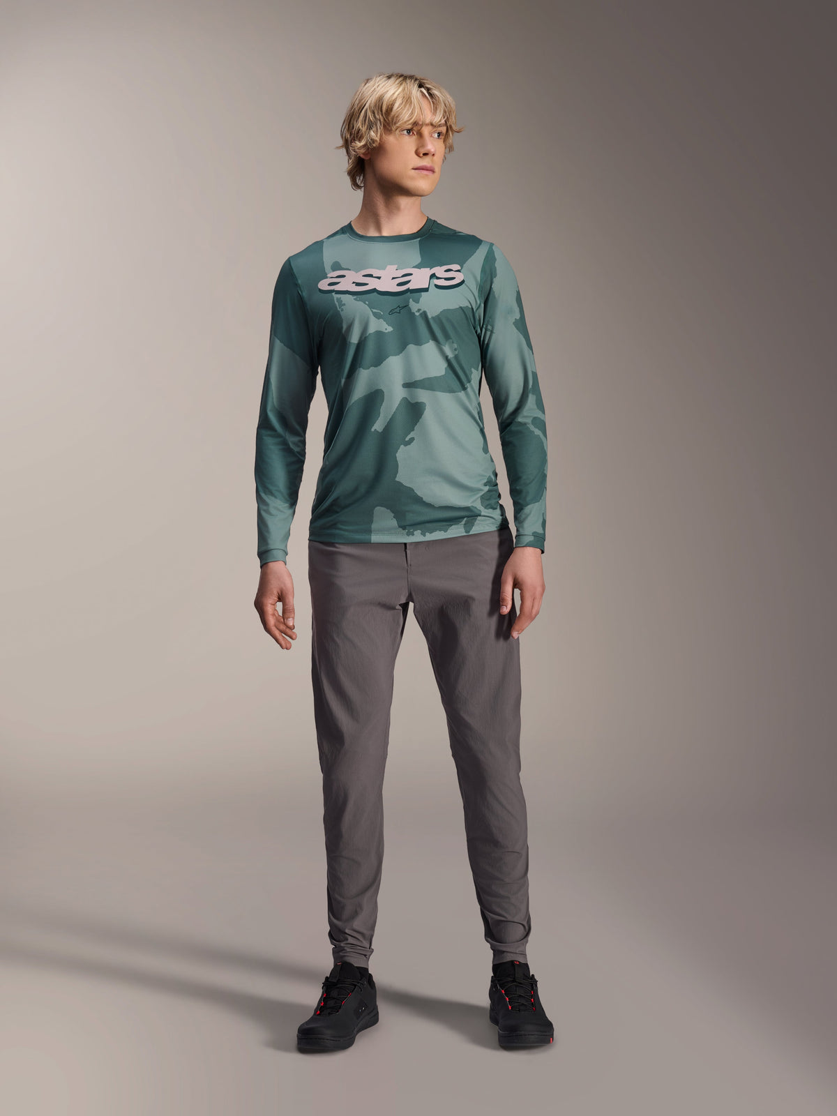 Alpinestars Maglia A-Dura Hollow Camo a maniche lunghe, Silver Pine Camo con motivo mimetico verde, grande logo "astars" bianco sul petto, indossato con pantaloni tecnici grigi e scarpe da ciclismo nere, abbigliamento MTB ad alte prestazioni
