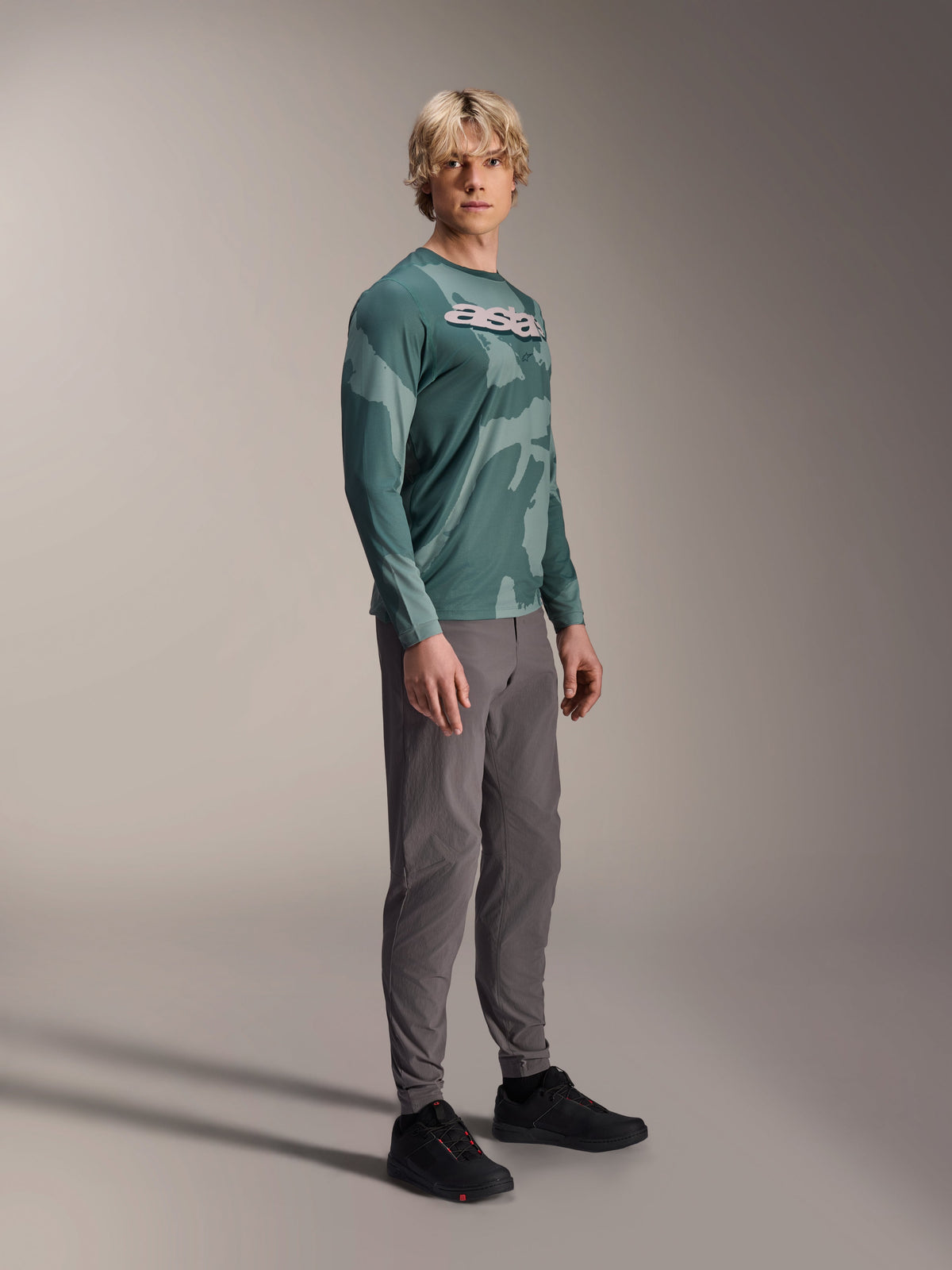 Maglia a maniche lunghe Alpinestars A-Dura Hollow Camo, Silver Pine Camo, indossata da un modello con pantaloni grigi e scarpe nere, con un motivo mimetico verde e un vistoso logo ASTARS bianco sul petto, abbigliamento MTB ad alte prestazioni