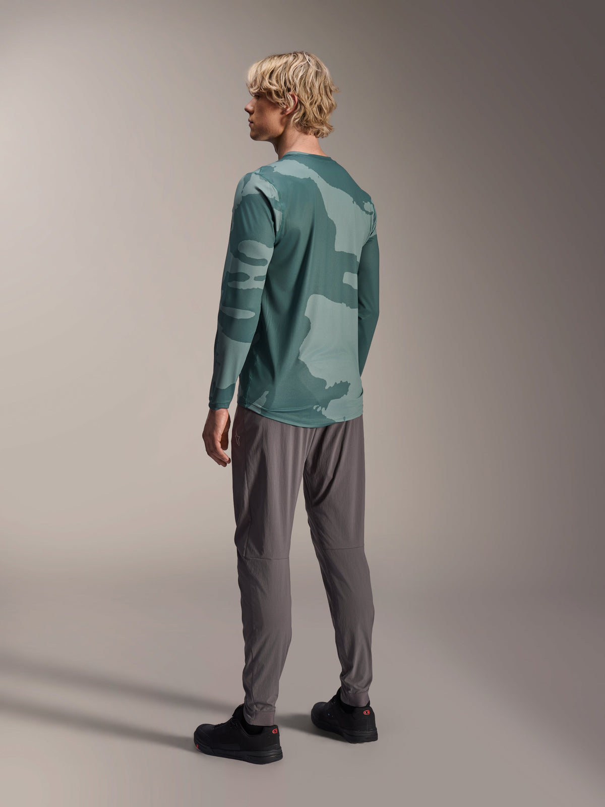 Maglia a maniche lunghe Alpinestars A-Dura Hollow Camo, Silver Pine Camo, vista posteriore del modello che indossa maglia mimetica verde con pantaloni grigi e scarpe nere, mostra il design a coda lunga e il motivo mimetico, abbigliamento tecnico da mountain bike
