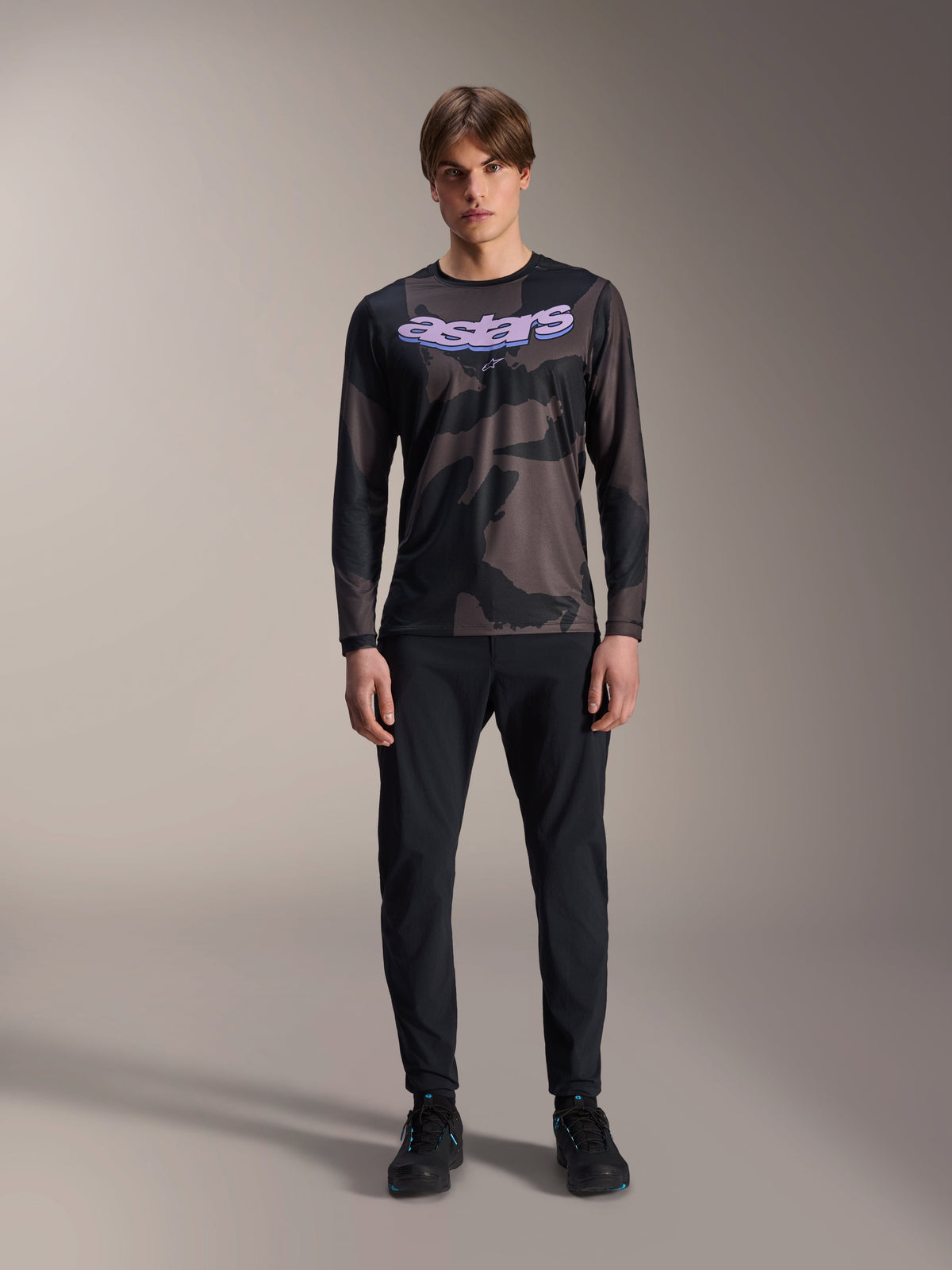 Modello che indossa la maglia a maniche lunghe Alpinestars A-Dura Hollow Camo in Black Camo, con logo 'astars' viola sul petto, abbinata a pantaloni neri e scarpe da ciclismo nere, abbigliamento per mountain bike