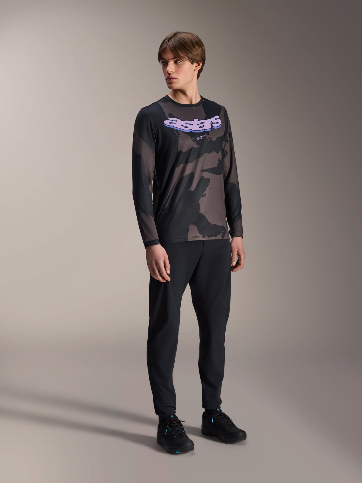 Modello che indossa la maglia a maniche lunghe Alpinestars A-Dura Hollow Camo, motivo Black Camo con grafica 'astars' viola sul petto, abbinata a pantaloni tecnici neri e scarpe nere, abbigliamento casual da MTB