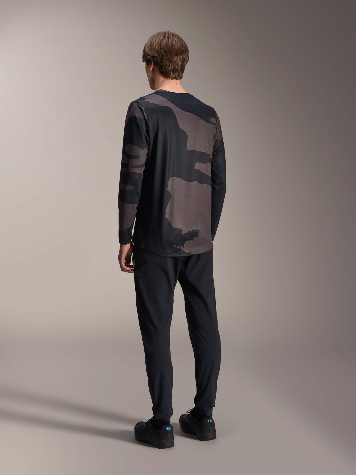 Alpinestars Maglia a maniche lunghe A-Dura Hollow Camo, Black Camo, vista posteriore del modello, maglia tecnica da mountain bike con orlo posteriore allungato, abbinata a pantaloni neri e scarpe da ciclismo, abbigliamento per prestazioni off-road dallo stile casual
