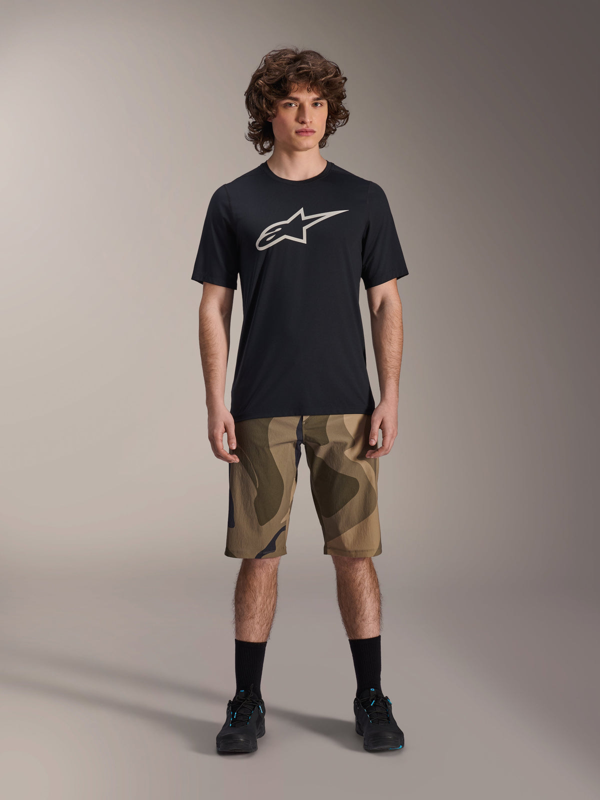 Alpinestars Maglia A-Dura Astar a maniche corte, nera con grande logo Astars bianco sul petto, indossata da un modello con pantaloncini MTB mimetici e scarpe da ciclismo nere, abbigliamento casual da mountain bike