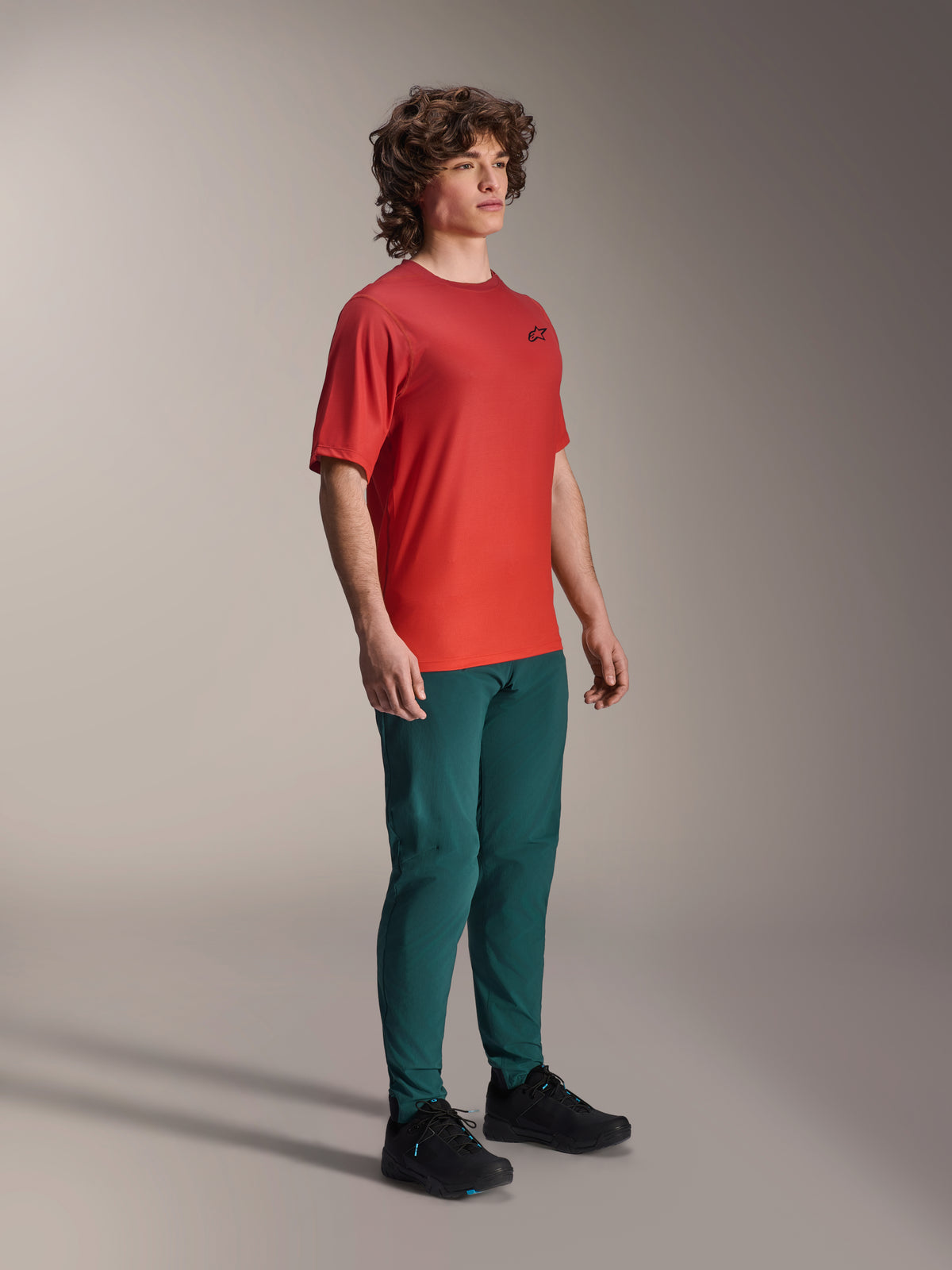 Modello che indossa i pantaloni Alpinestars A-Dura in Dark Pine, abbinati a una maglia a maniche corte A-Dura Rise rossa e scarpe da MTB nere, abbigliamento tecnico da mountain bike in studio