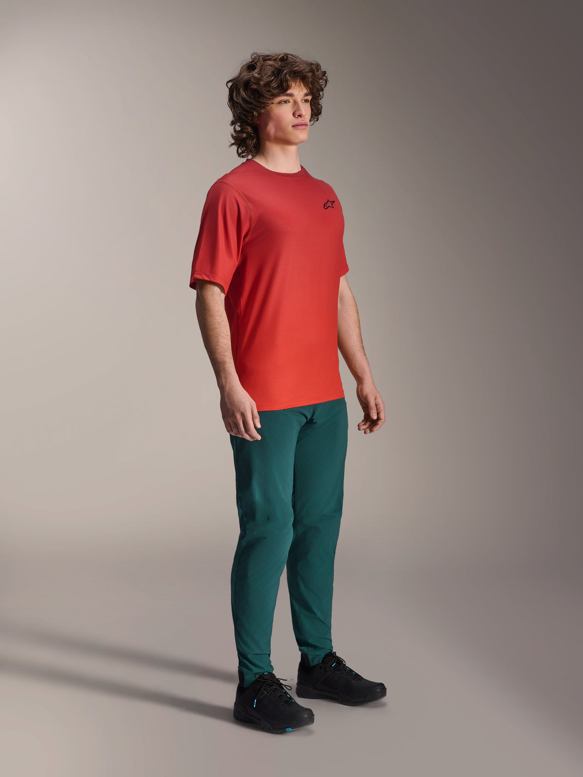 Alpinestars Maglia A-Dura Rise a maniche corte, Spicy Orange, con un piccolo logo nero sul petto, indossata con pantaloni verde petrolio scuro e scarpe nere, abbigliamento da mountain bike leggero e traspirante