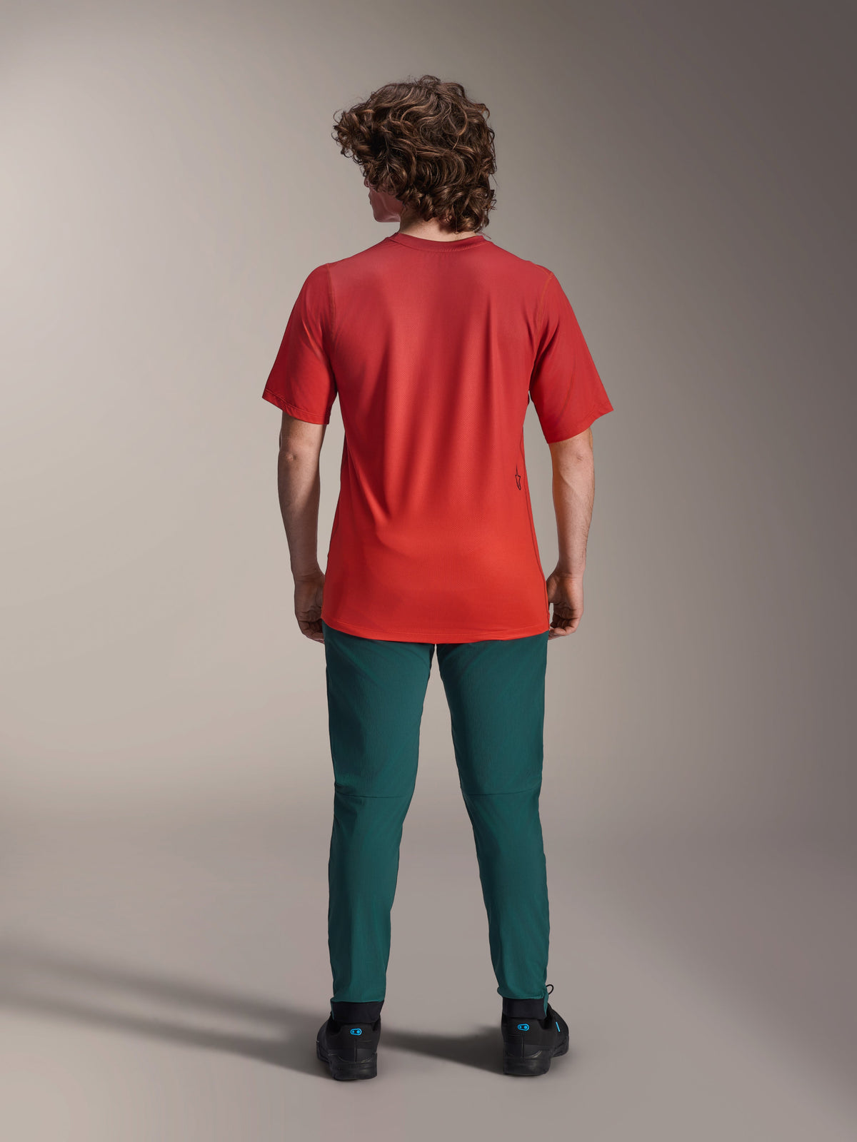 Maglia a maniche corte Alpinestars A-Dura Rise, Spicy Orange, vista posteriore su un modello che indossa pantaloni ottanio scuro e scarpe nere, con coda allungata e un piccolo logo serigrafato sul retro, abbigliamento tecnico leggero per MTB.