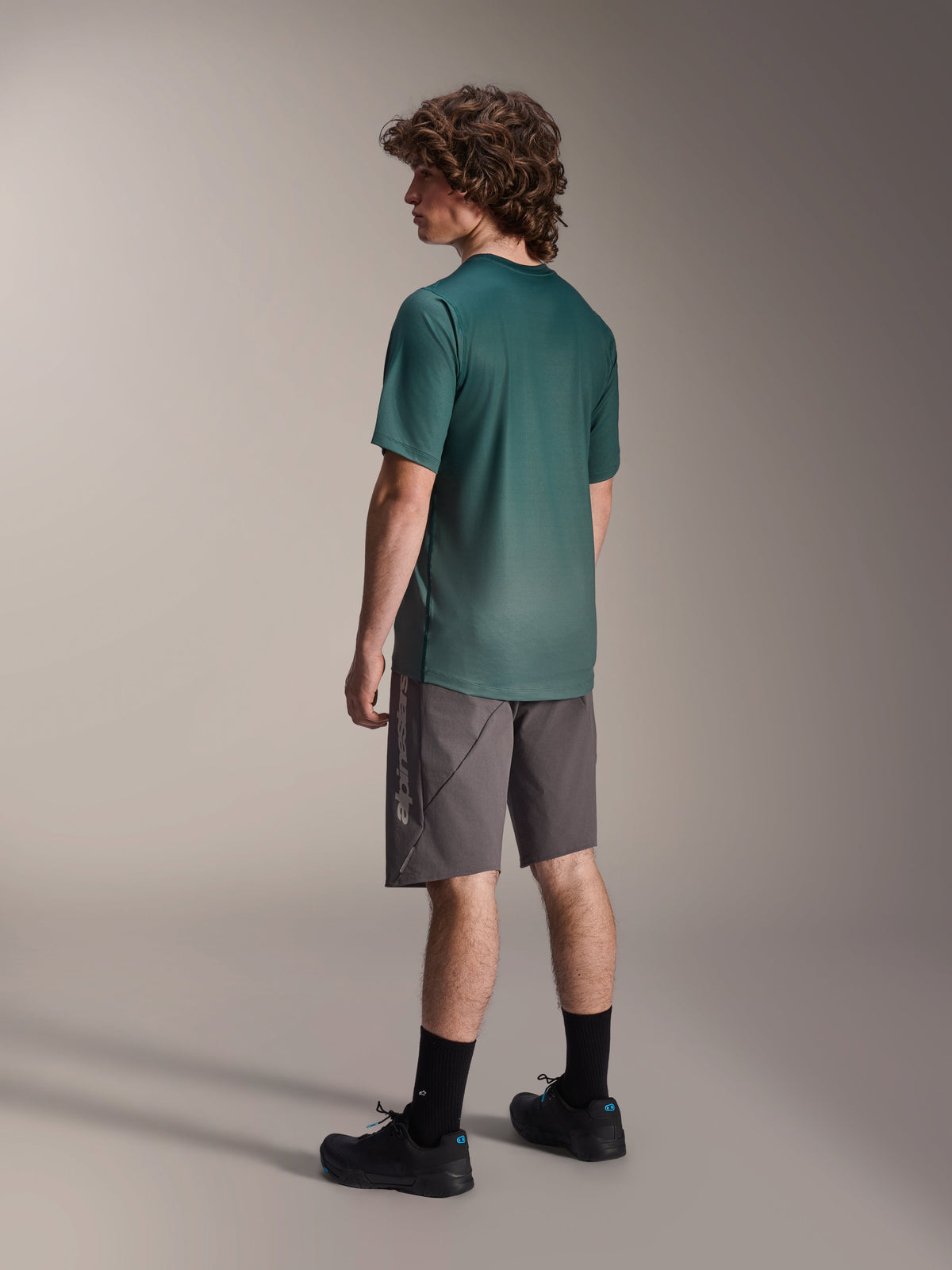Alpinestars A-Dura Rise Jersey, maglia MTB a maniche corte, Verde Silver Pine, indossata con pantaloncini grigi e scarpe nere, vista posteriore che mostra la coda più lunga e la vestibilità regolare, tessuto leggero traspirante per trail riding