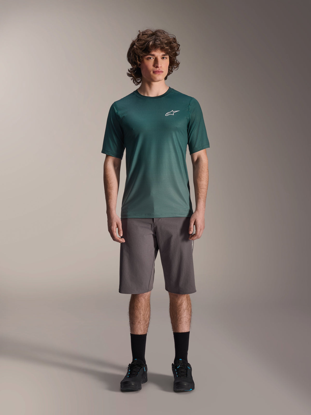 Modello che indossa la maglia a maniche corte Alpinestars A-Dura Rise in Silver Pine, maglia da ciclismo verde con piccolo logo bianco sul petto, abbinata a pantaloncini da mountain bike grigi e calzature nere, abbigliamento casual da trail MTB